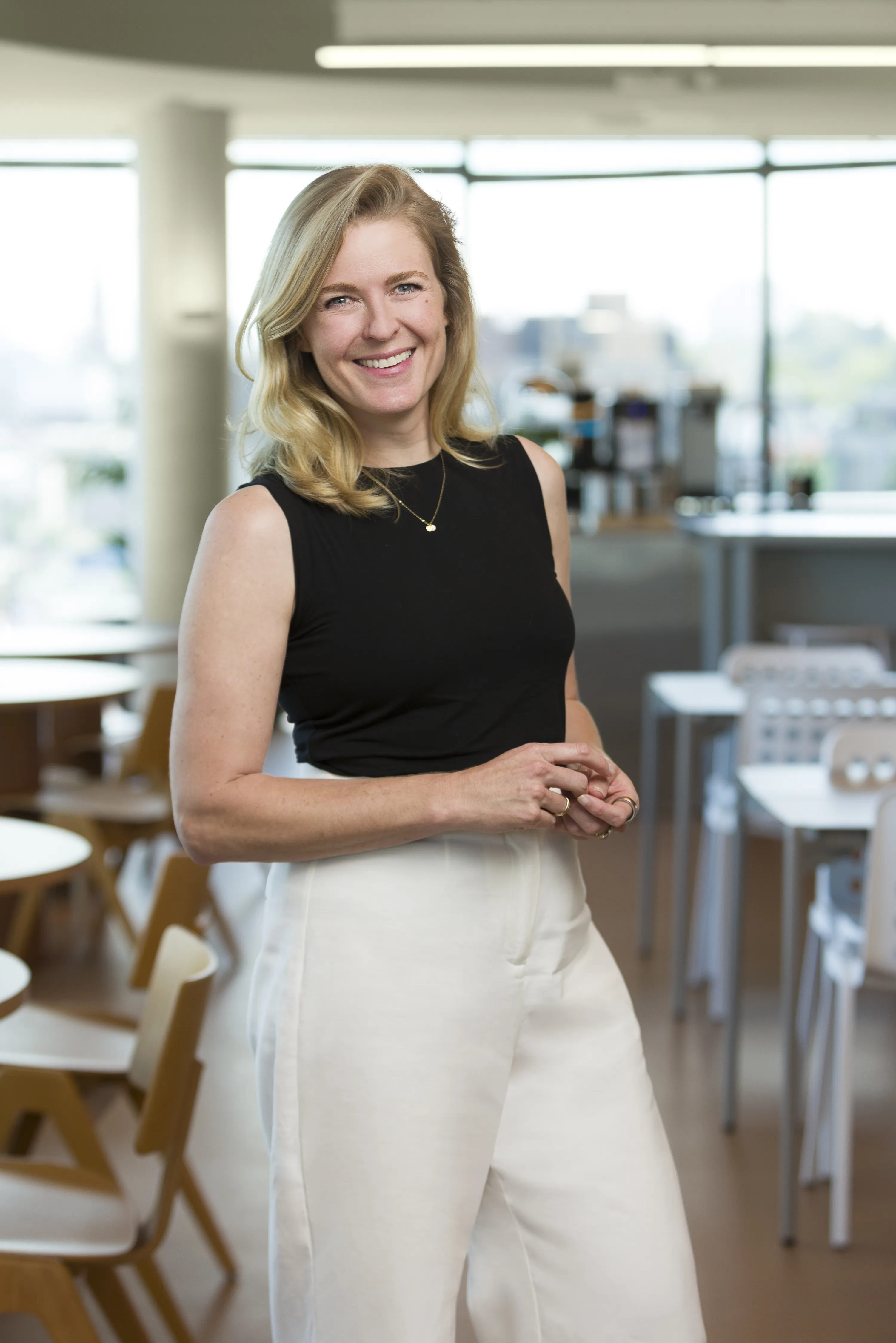 Kate Collinson, a smiling, blonde woman wearing a black sleeveless top and white pants standing indoors with tables and chairs in the background.