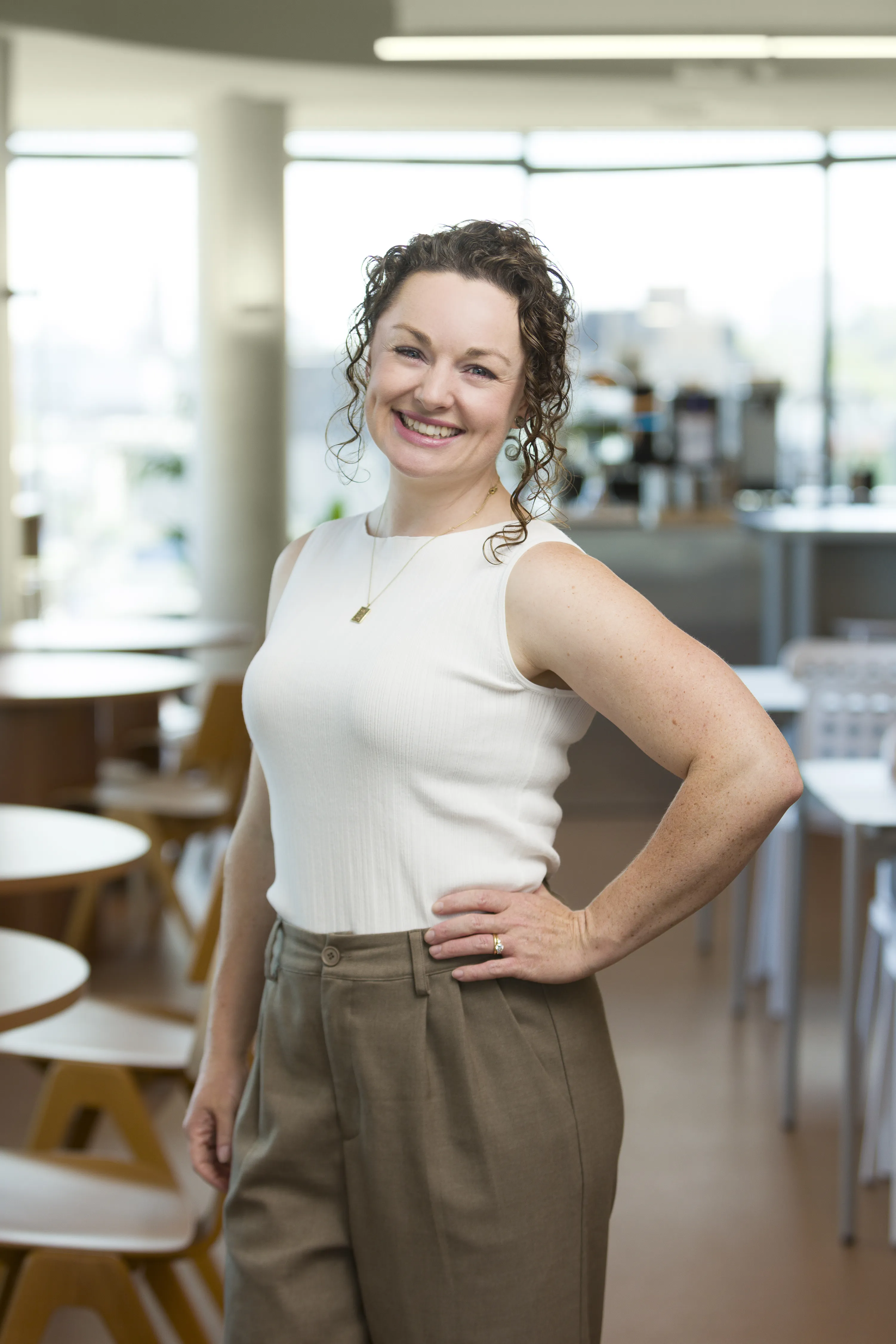 Melanie Gray, a smiling woman with curly brown hair wearing a sleeveless white top and brown pants standing with hand on hip in a bright office space.