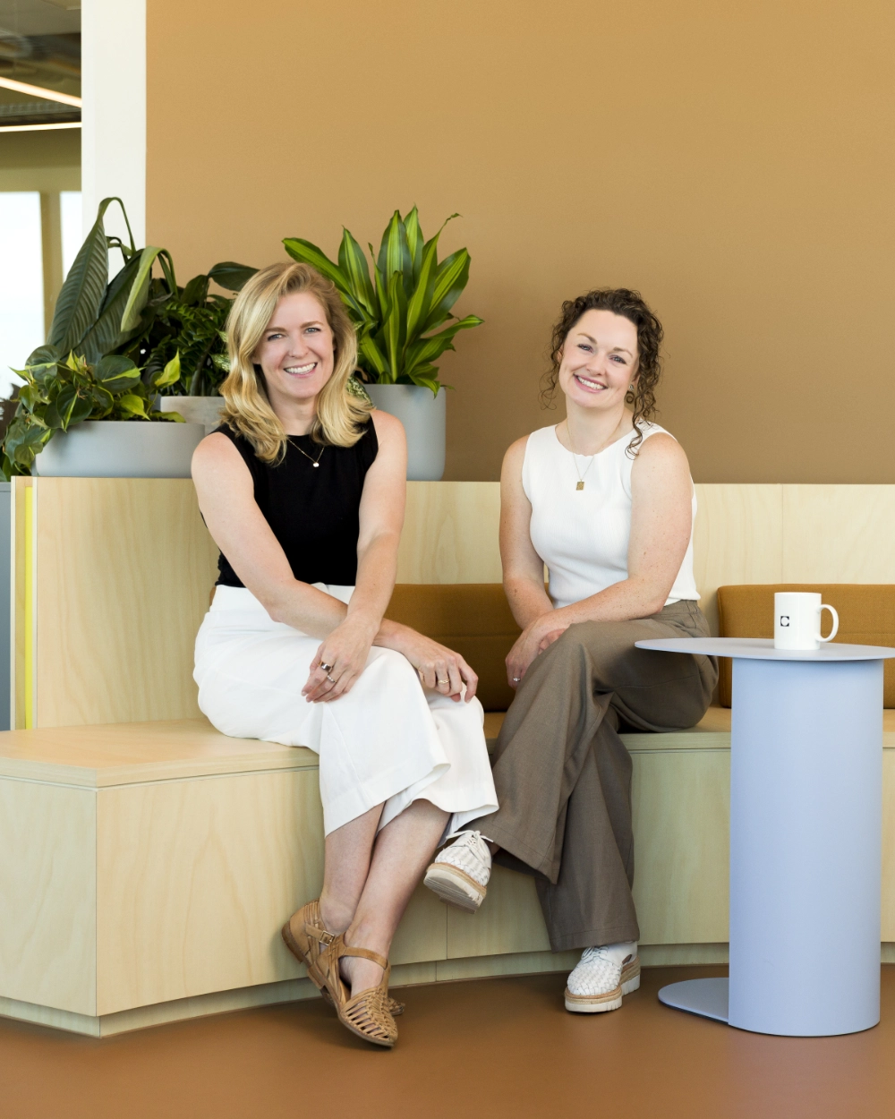 Kate and Mel are two women smiling while seated on a light wood bench with green plants in the background and a small white table with a mug beside them.