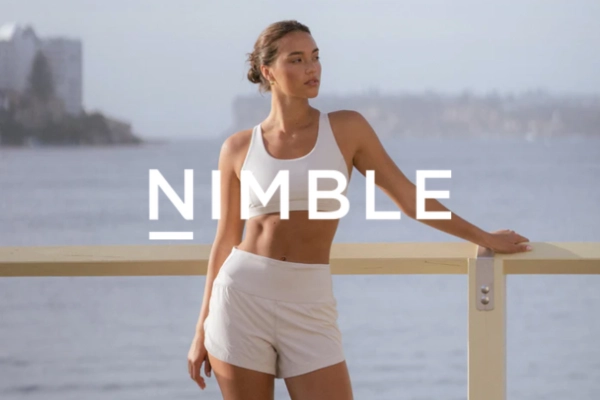 Woman in white activewear posing outdoors by a railing with water and cityscape in the background and the text 'NIMBLE' overlayed.