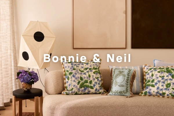 Cozy beige sofa with floral and embroidered cushions, next to a modern lamp and a small table with a flower vase, text 'Bonnie & Neil' overlaid.