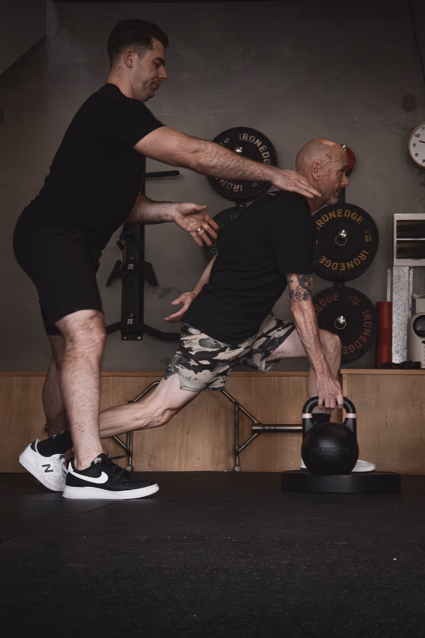 Trainer guides an older man doing a kettlebell lunge exercise in a gym setting.
