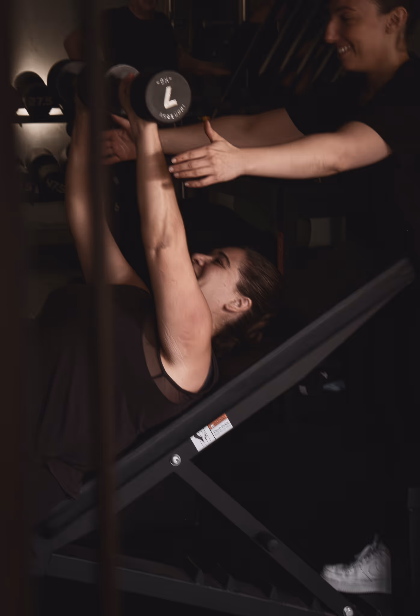 Woman lying on a bench lifting a dumbbell with assistance from a smiling trainer in a dim gym.