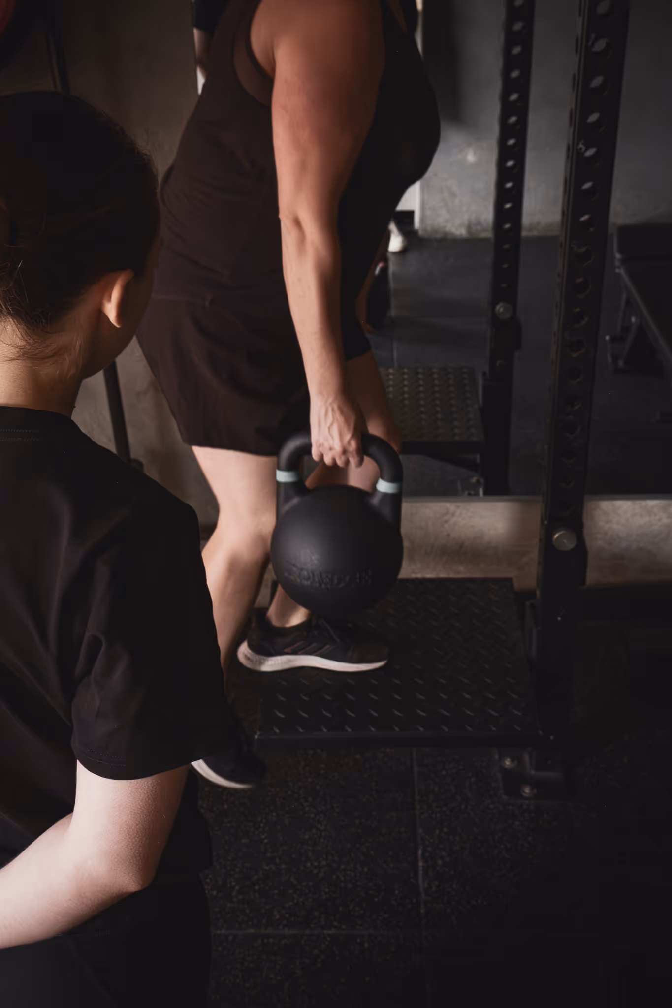 Person lifting a black kettlebell on a gym platform with another person watching nearby.