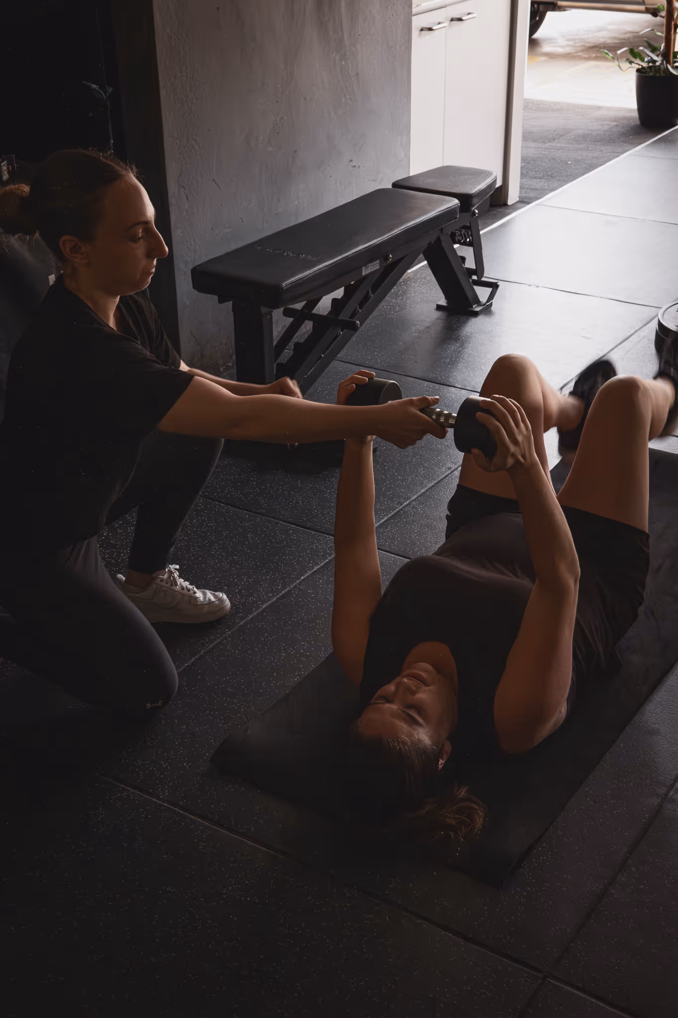Trainer assists woman lying on mat while lifting dumbbell in gym setting.