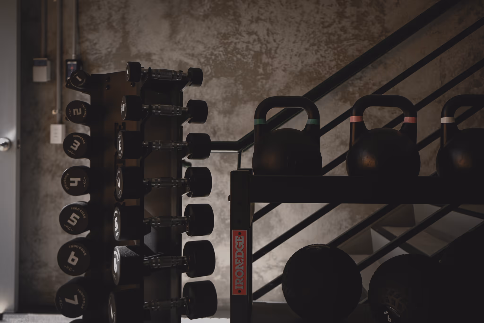 Dumbbells and kettlebells stacked on racks in a dimly lit gym with concrete walls and metal stairs.