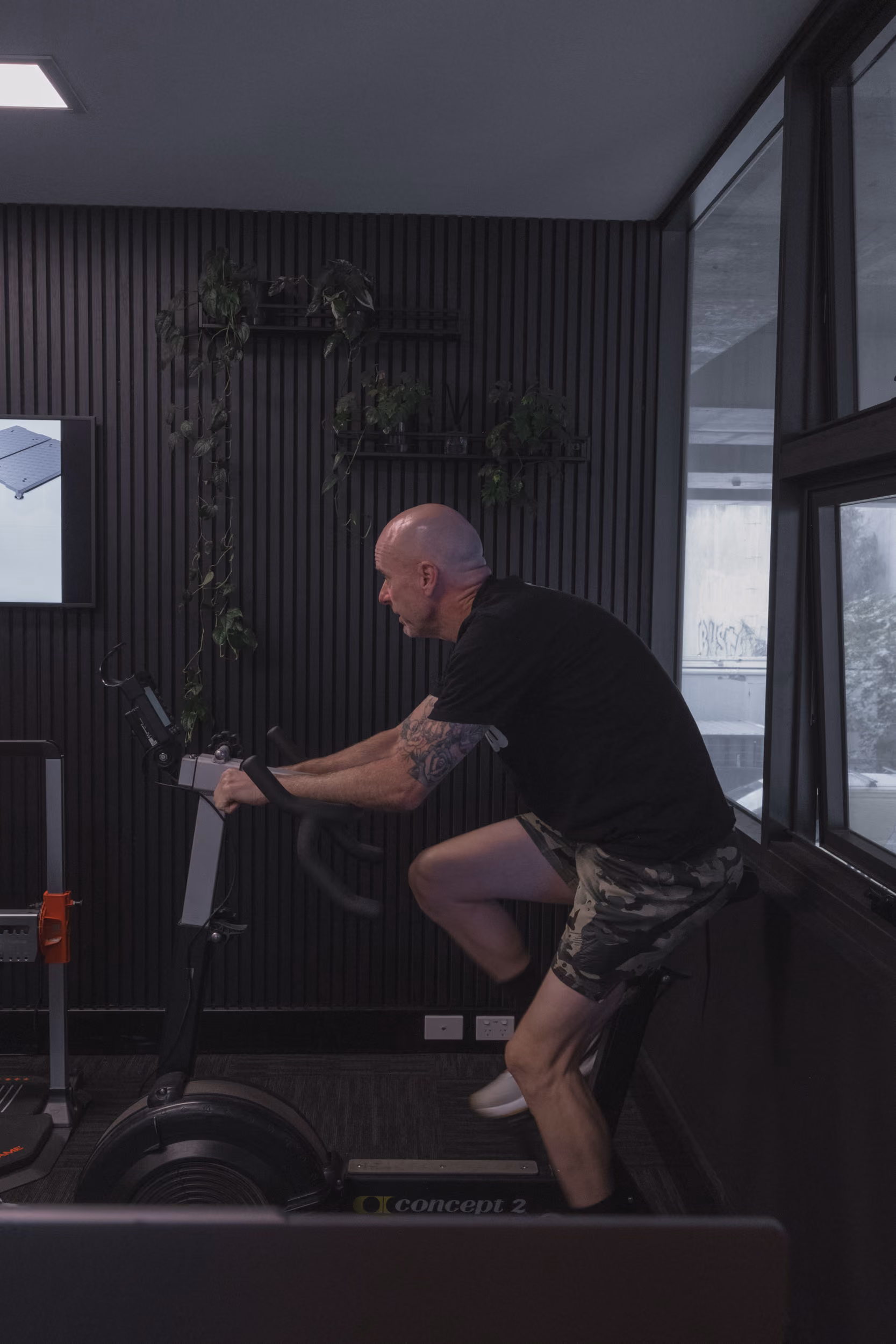 Bald man with tattoos in black t-shirt and camo shorts exercising on a stationary bike indoors.