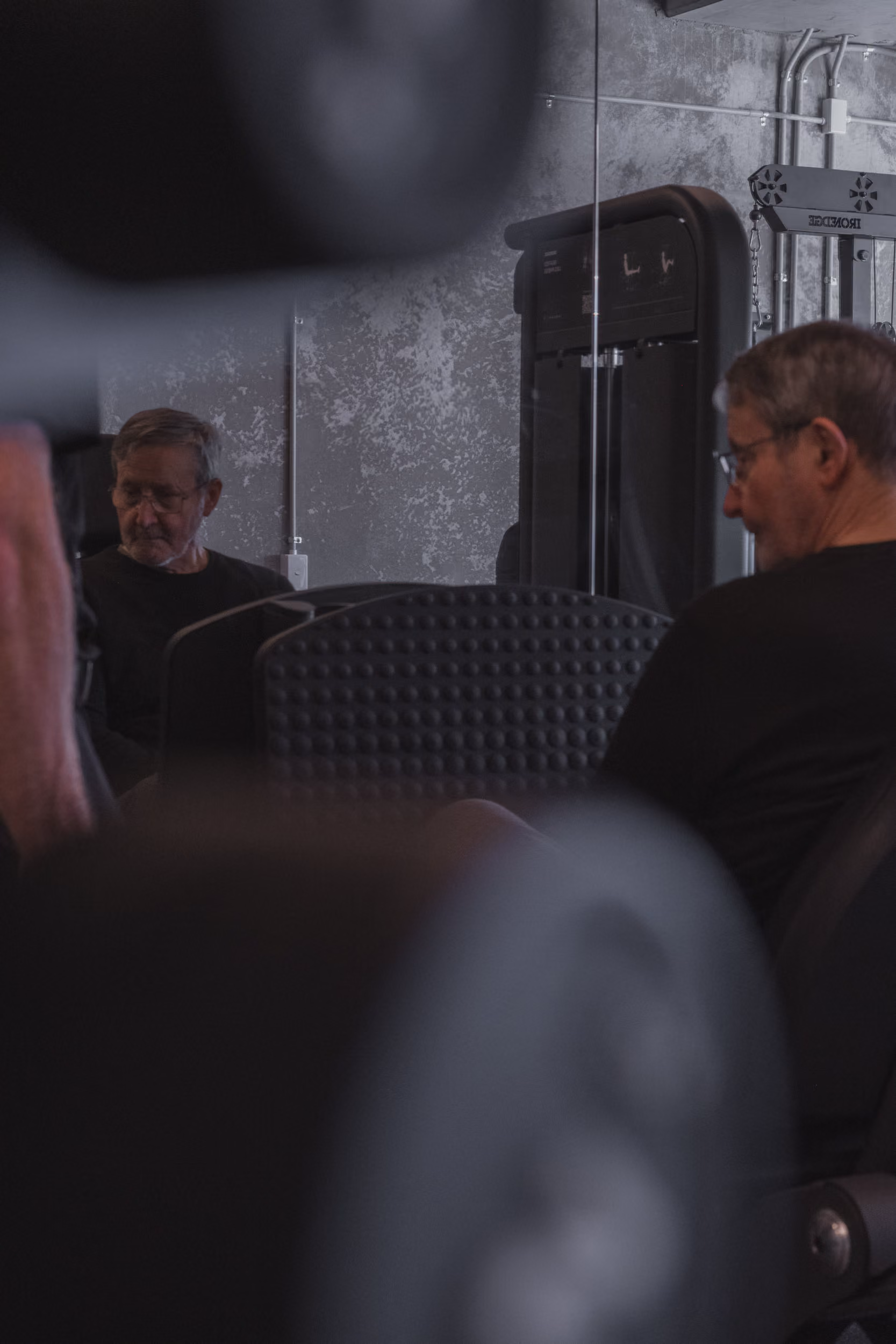 Older man in glasses sitting in gym surrounded by blurred weights and workout equipment.