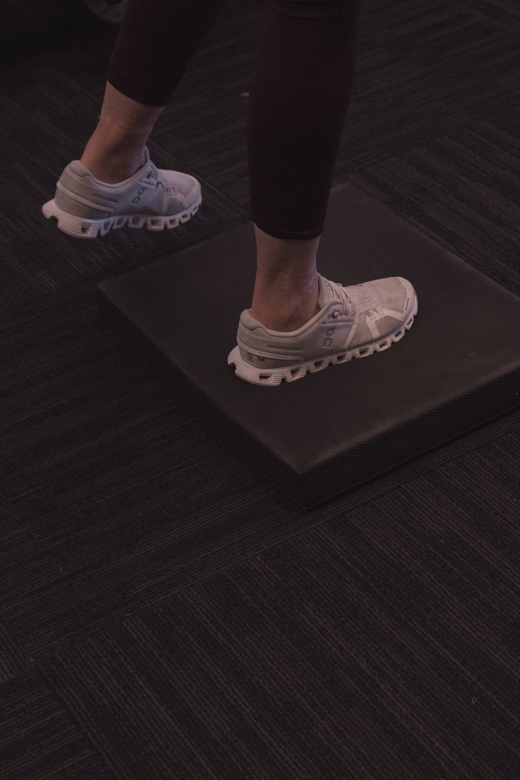 Person wearing white sneakers balancing one foot on a black cushion on a dark carpeted floor.
