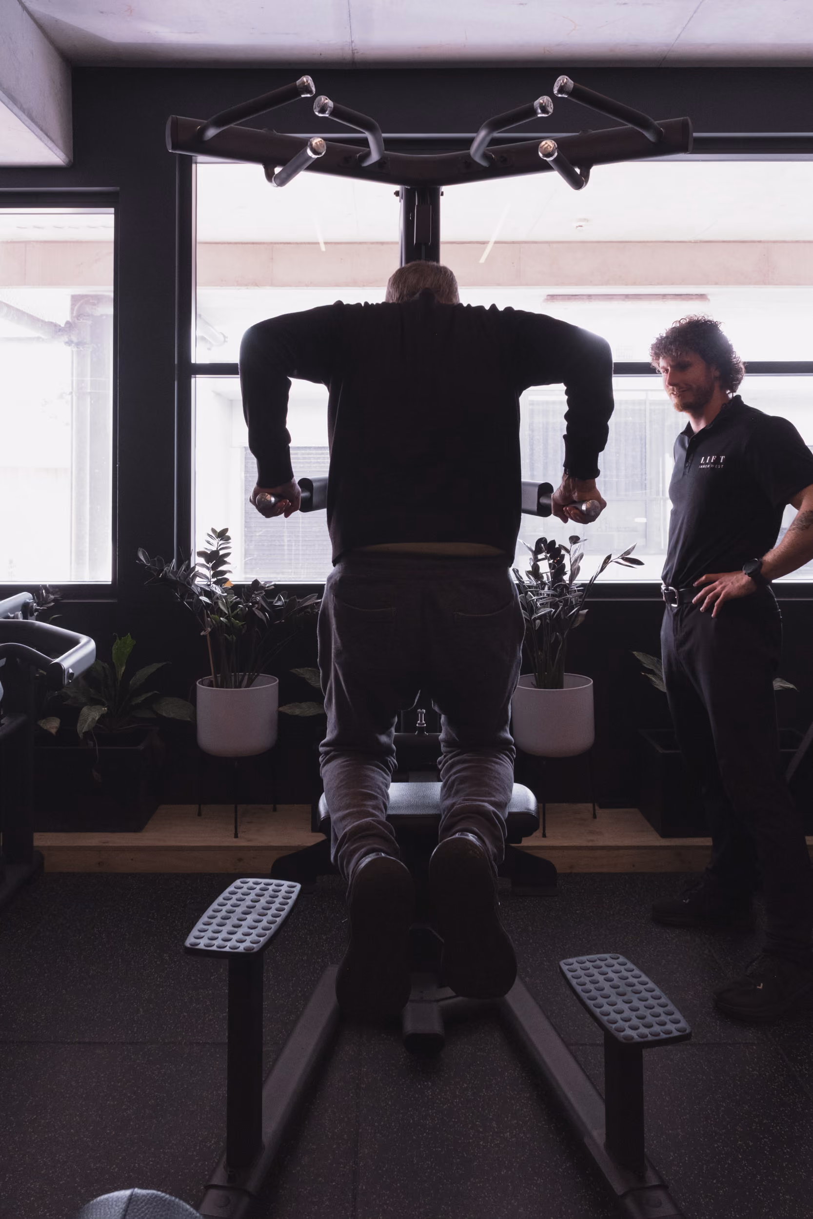 Person doing assisted dips on parallel bars in gym with trainer watching nearby.
