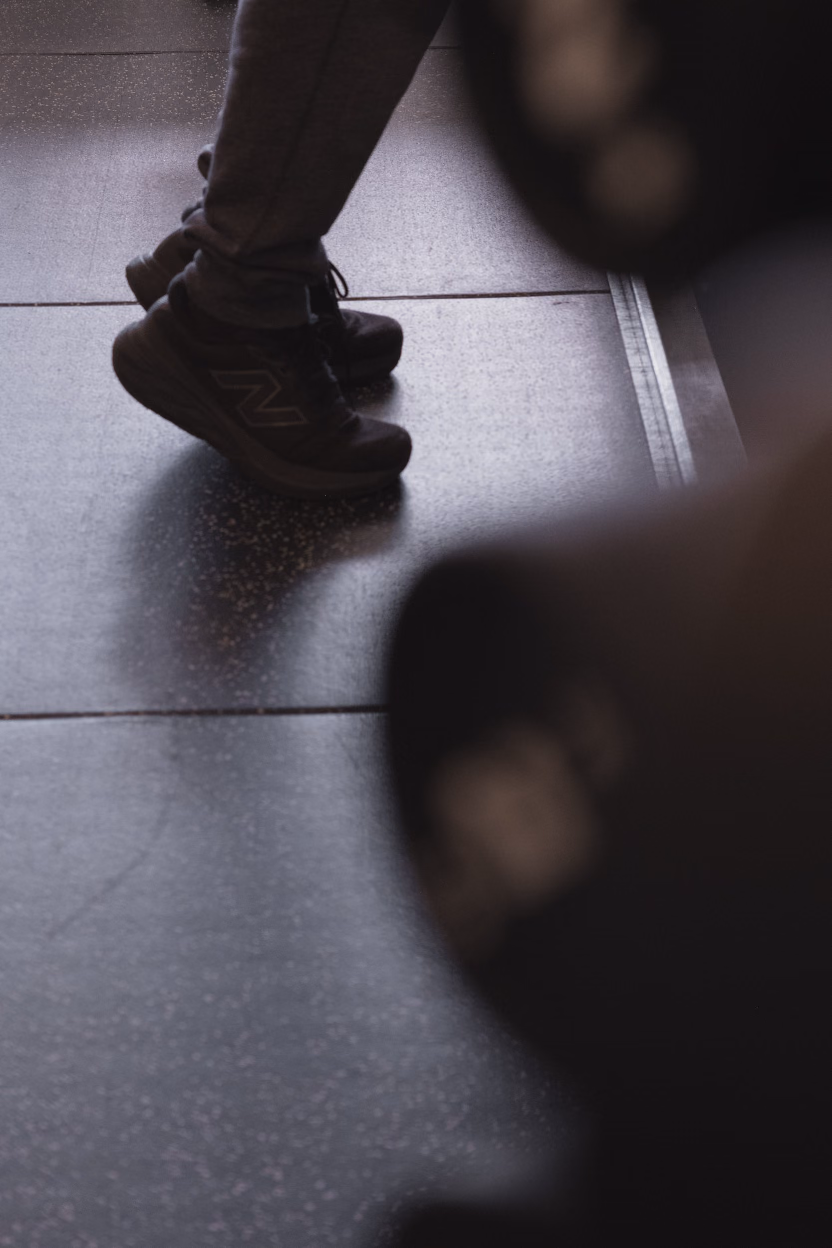 Person wearing dark New Balance shoes standing on a gym floor next to a blurred dumbbell.