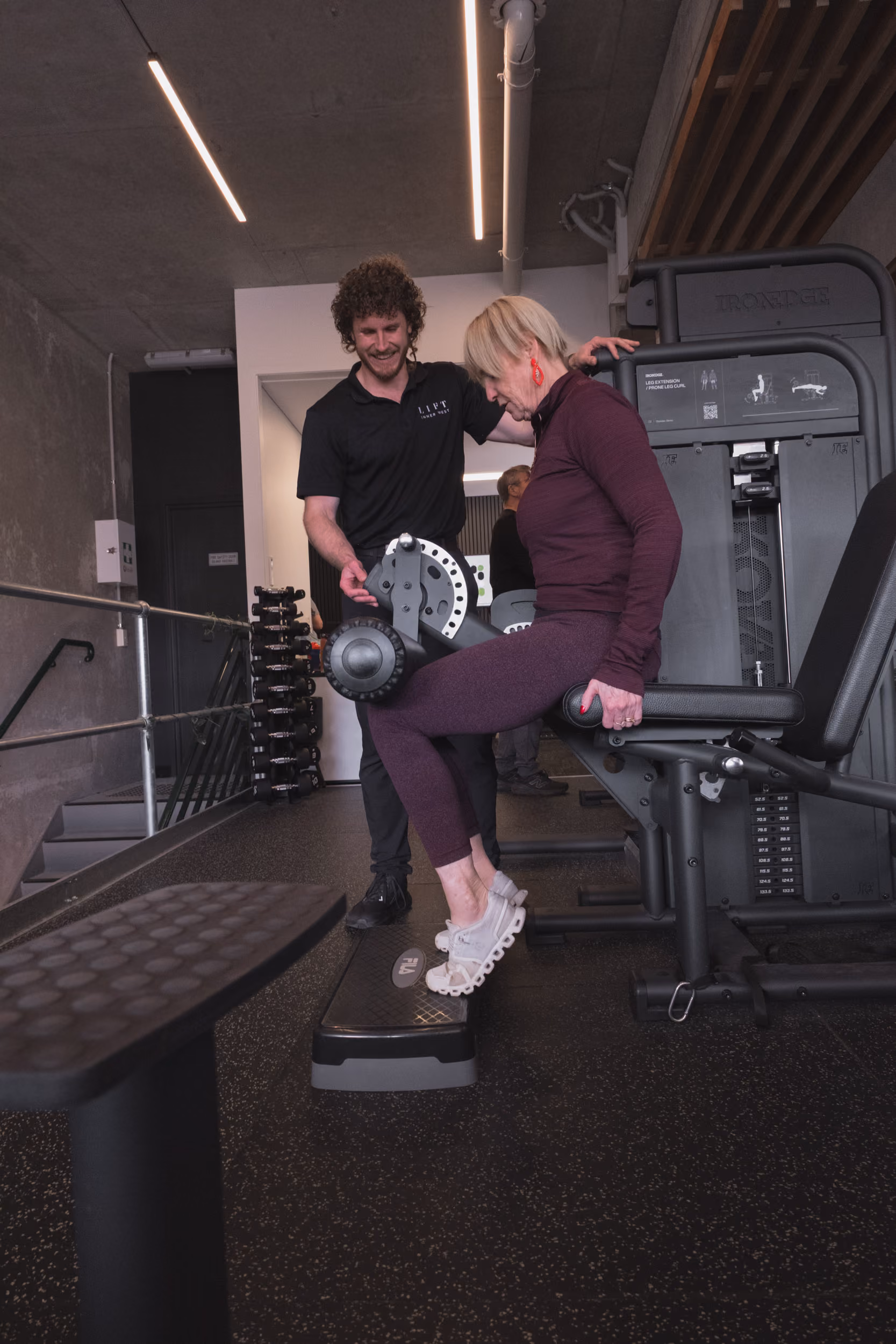 Trainer assists woman doing leg curls on exercise machine in a gym setting.
