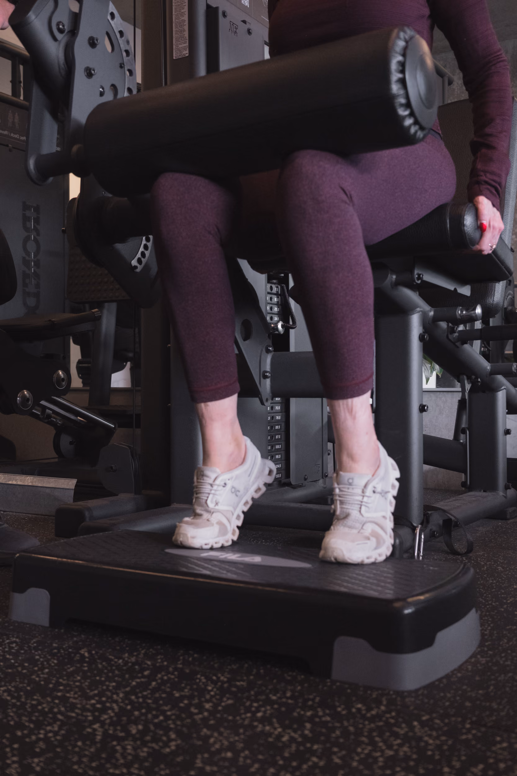 Person wearing purple leggings using a leg curl machine in a gym with white athletic shoes.