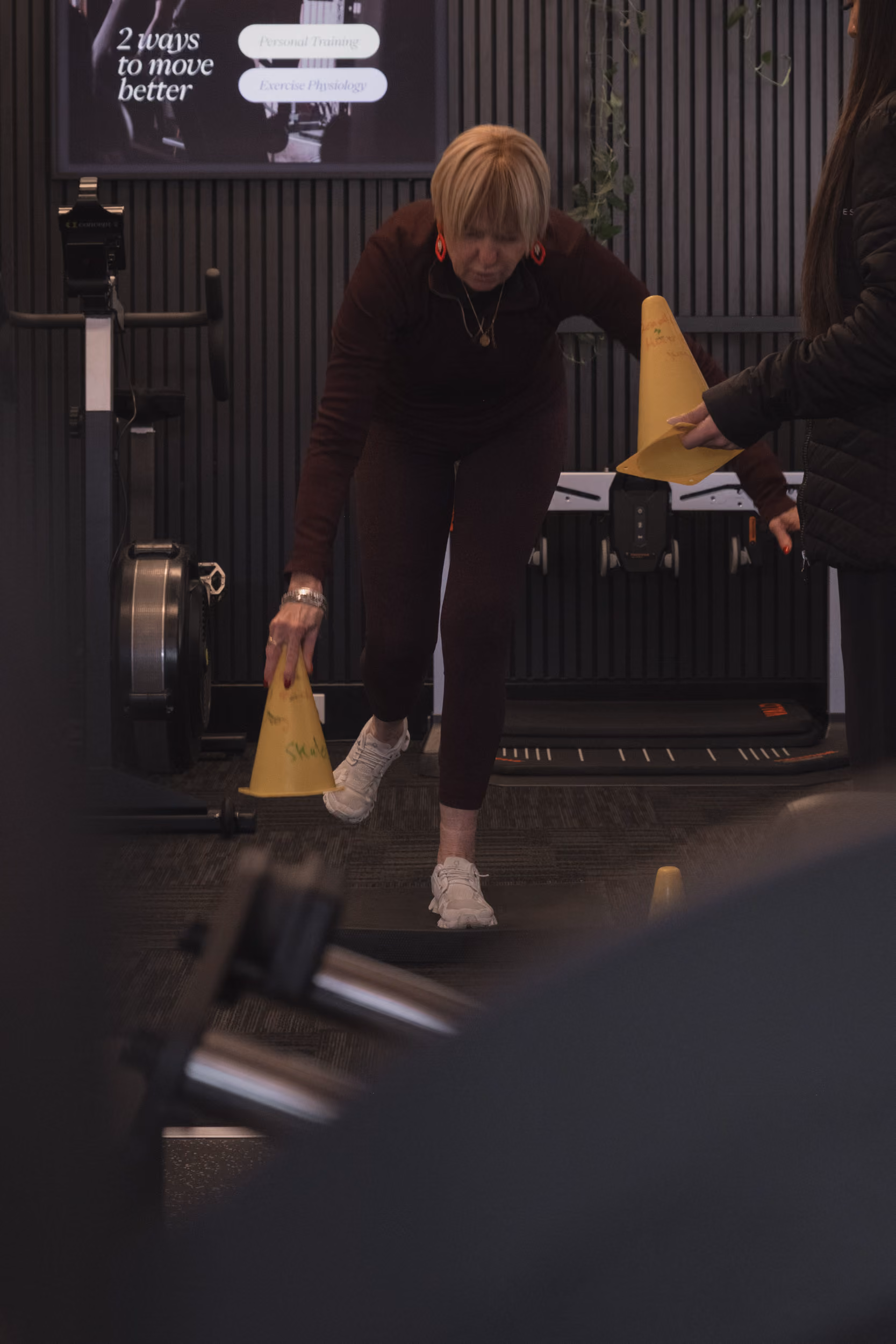 Older woman balancing on one leg while placing a yellow cone on the floor in a gym.