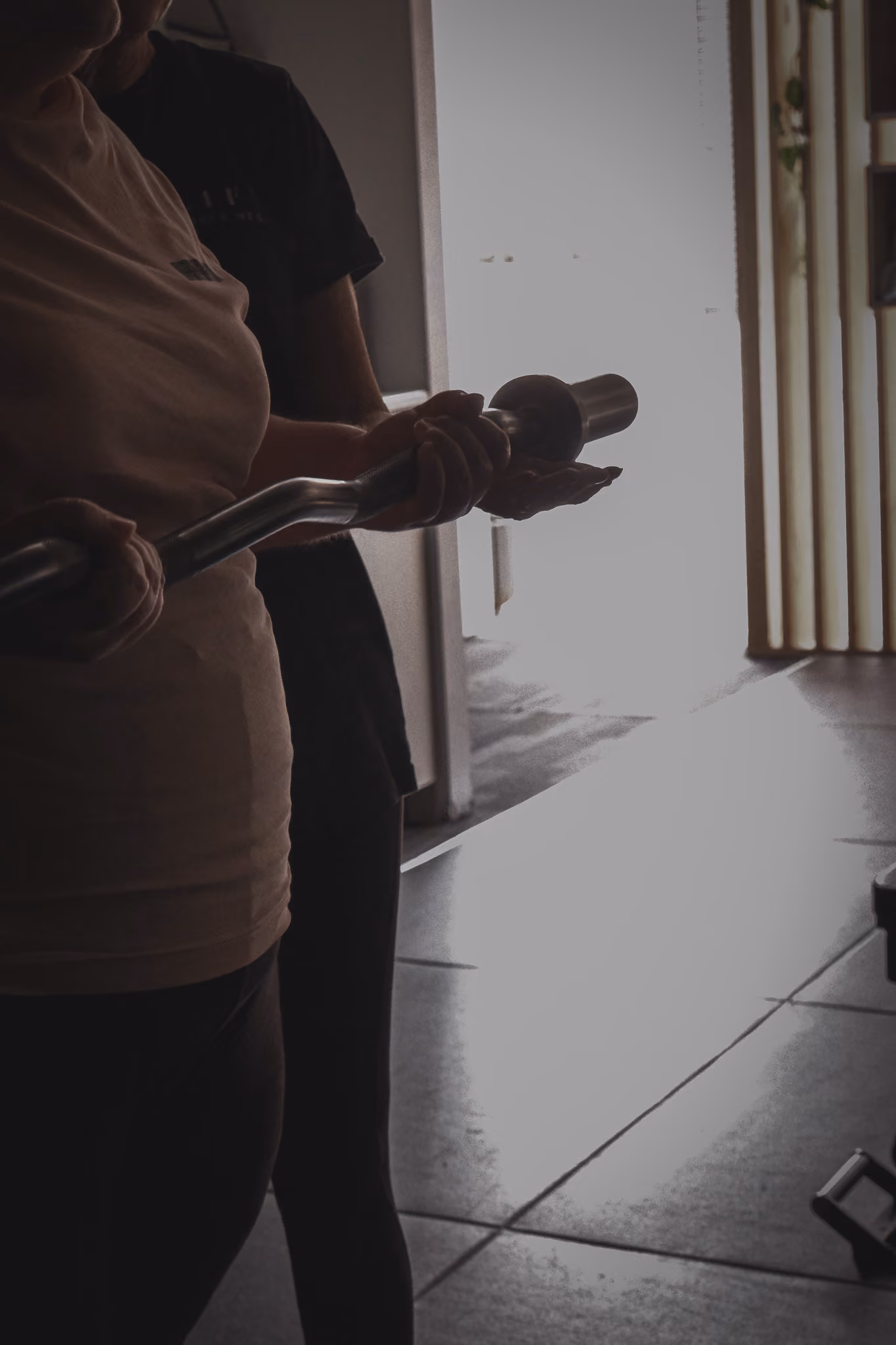 Two people holding a metal bar together in a dimly lit gym or workout area.