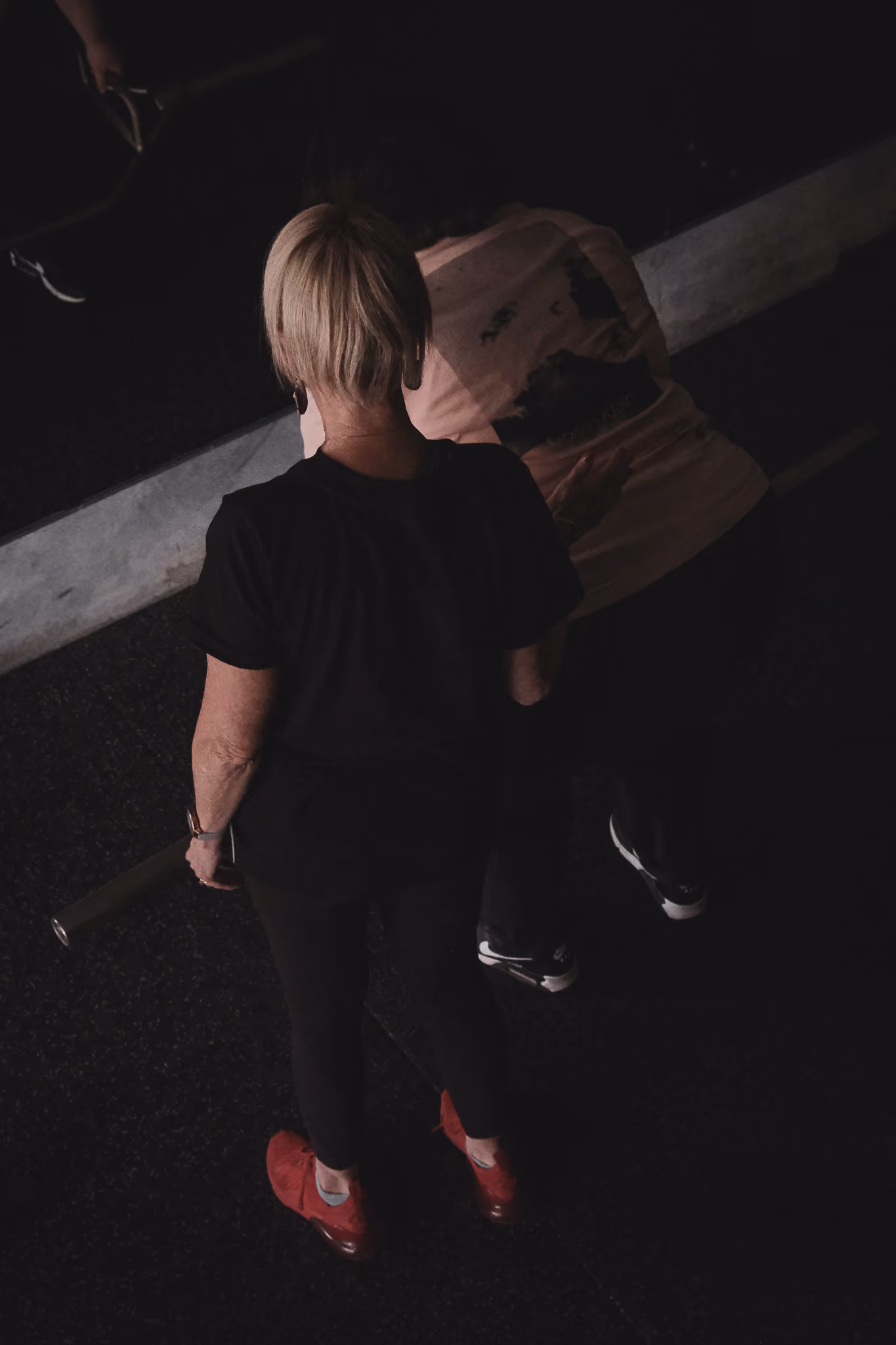 Two people from above in dim light; one stands with red shoes and the other leans forward.