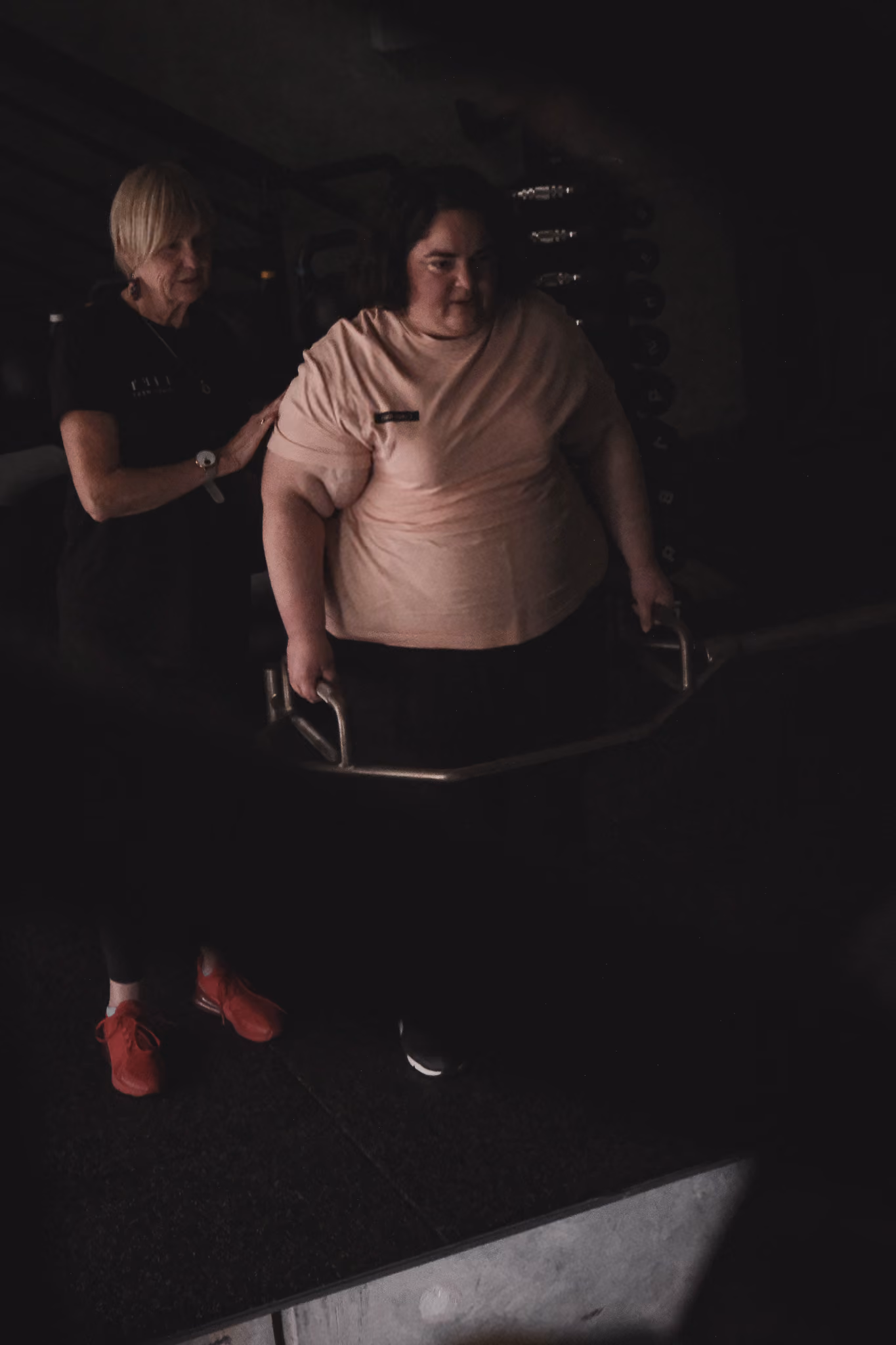 Person lifting barbell with trainer's support in a dimly lit gym setting.
