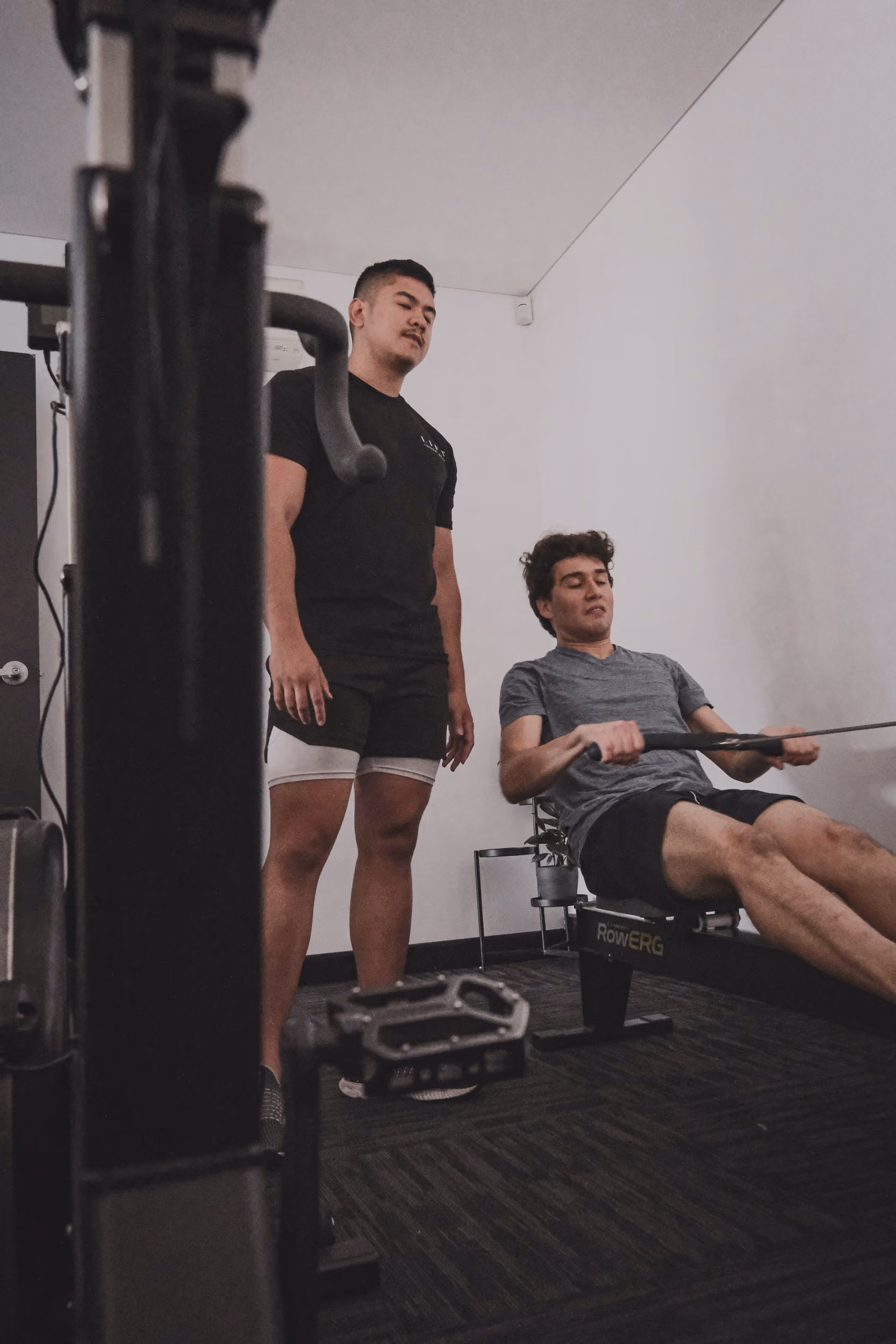 Man exercising on rowing machine with another man standing beside him watching.