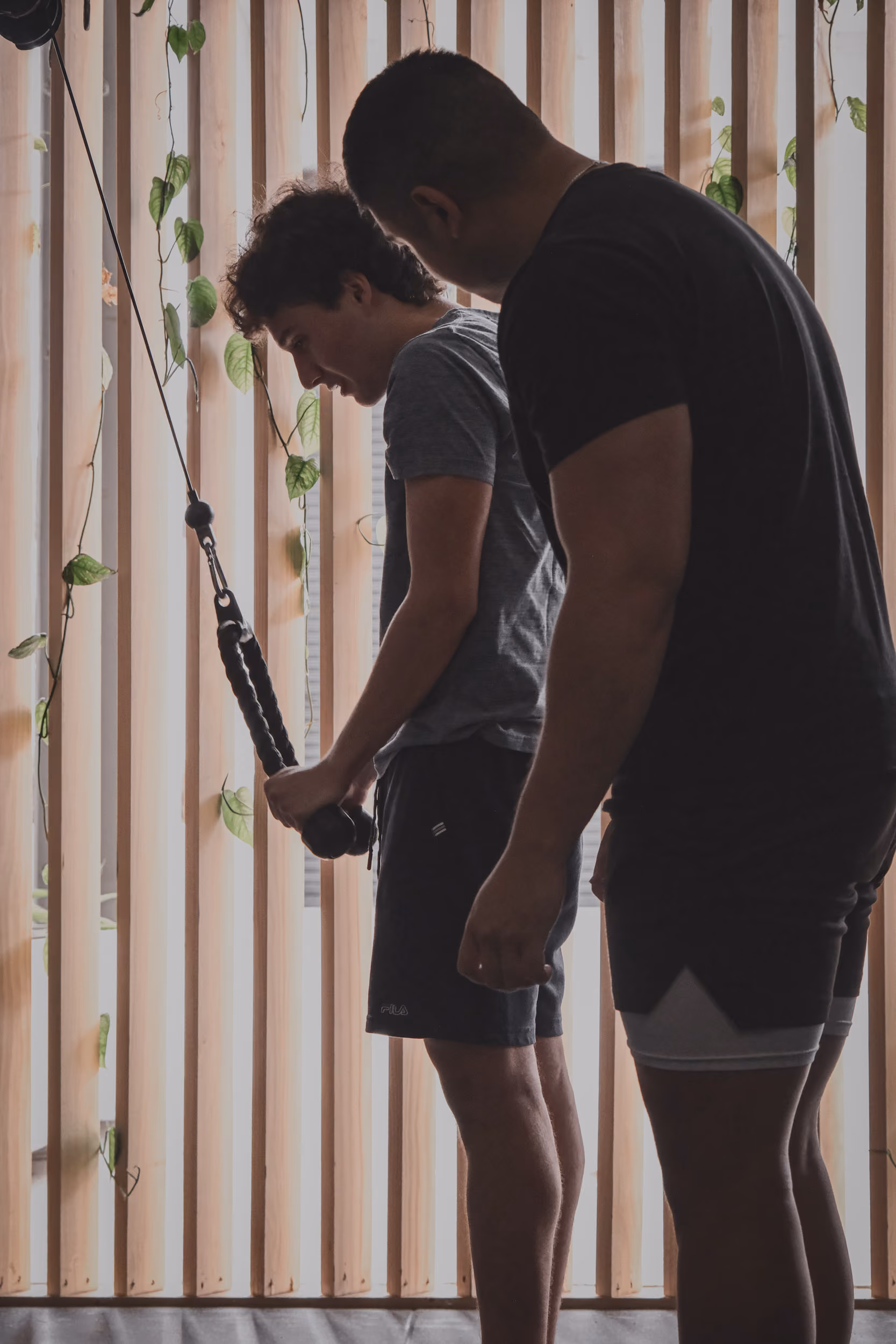 Two men standing near a wooden slat wall with vines, one holding gym cable equipment.