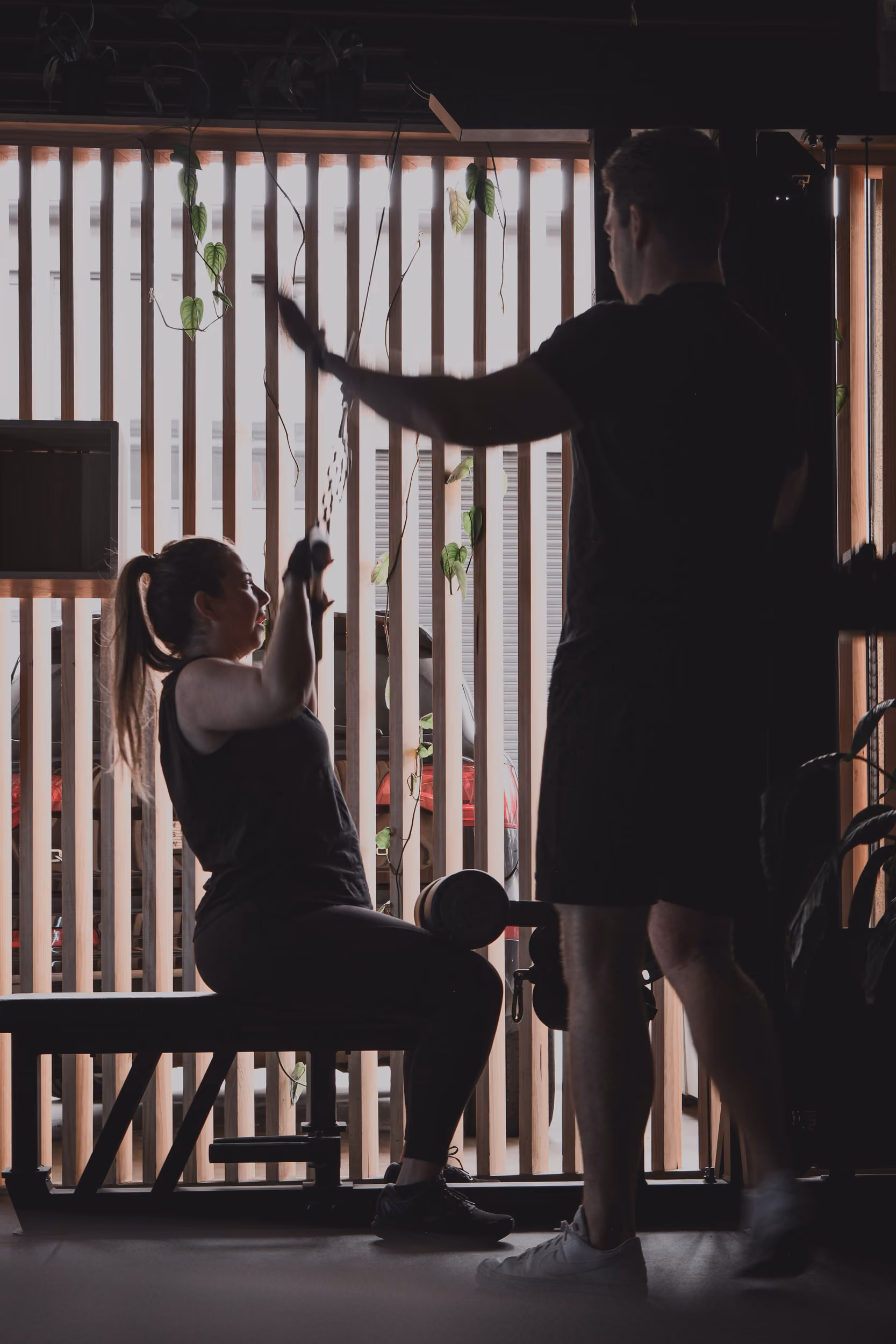 Woman on exercise bench pulling cable with a trainer supervising in a gym with wooden slats and plants.