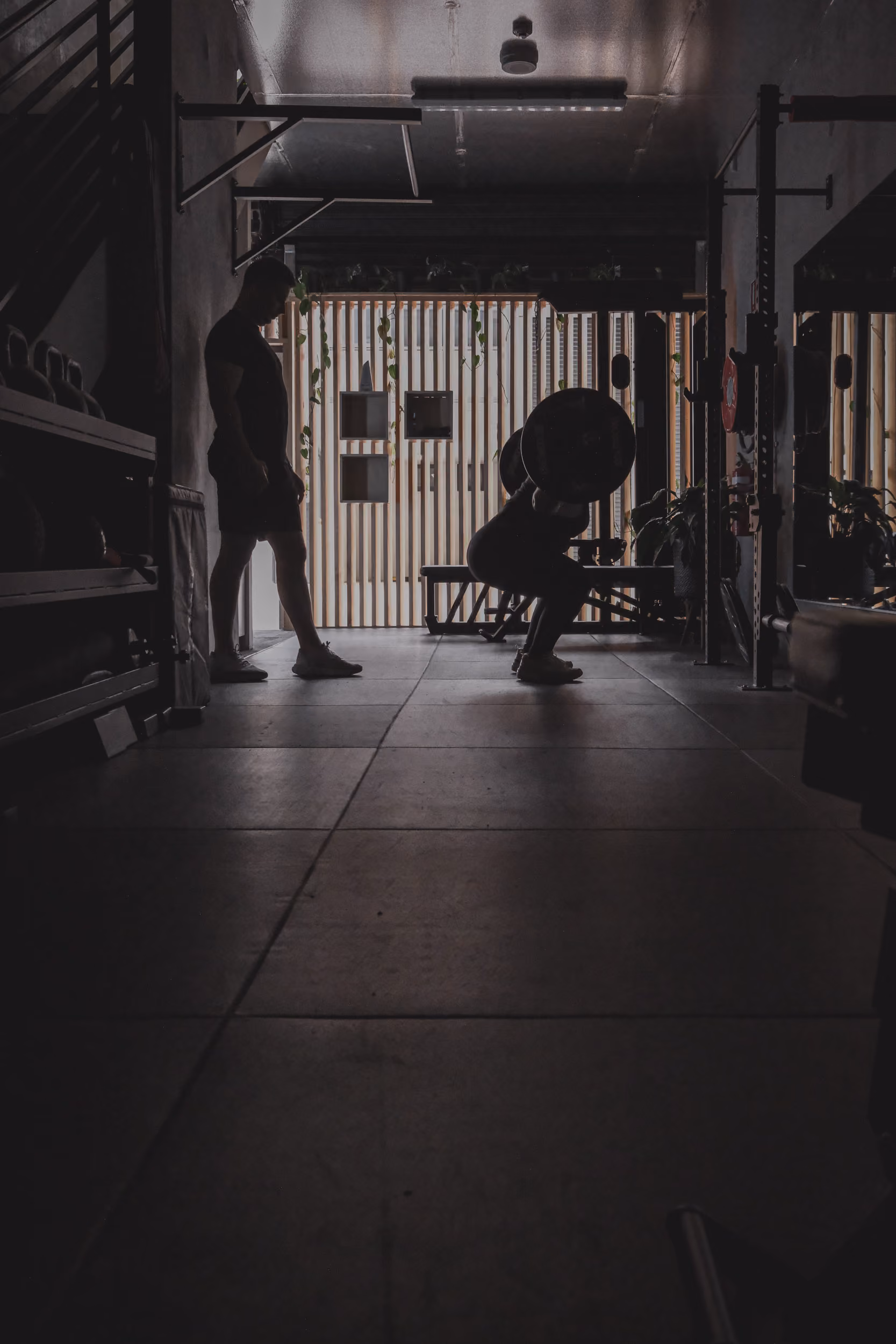 Silhouettes of two men in a gym, one squatting with a barbell while the other watches nearby.
