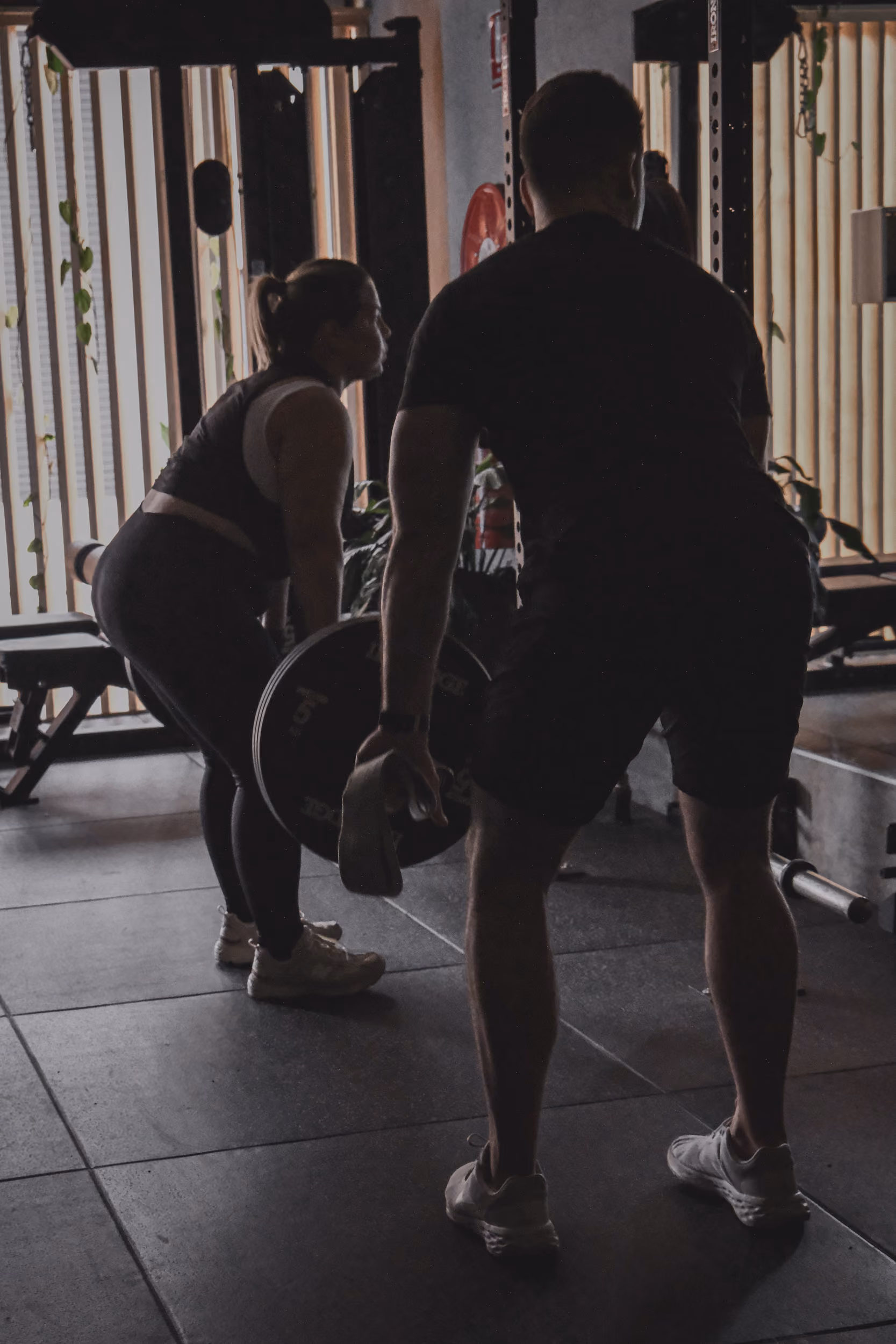 Man coaching woman lifting a weighted barbell in a gym with natural light through windows.
