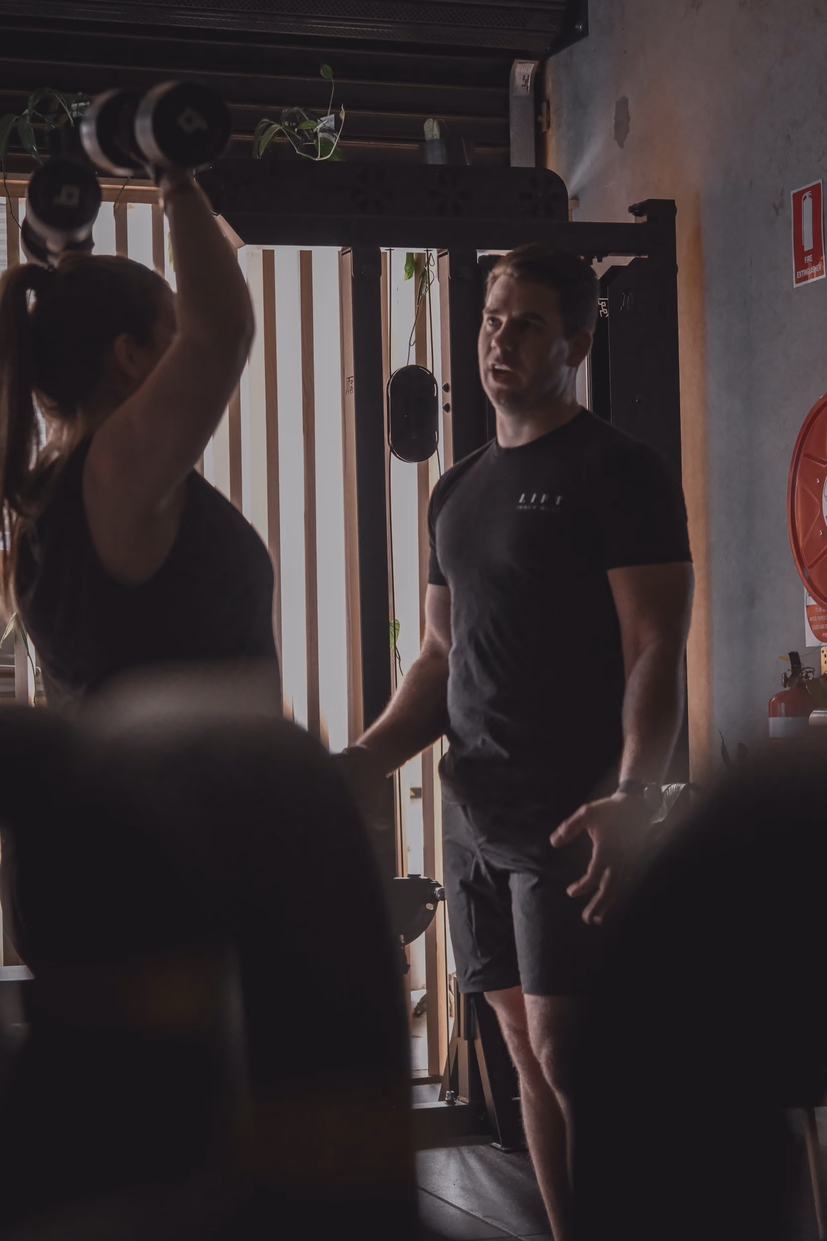 Woman lifting dumbbells overhead while a male trainer coaches her in a gym setting.