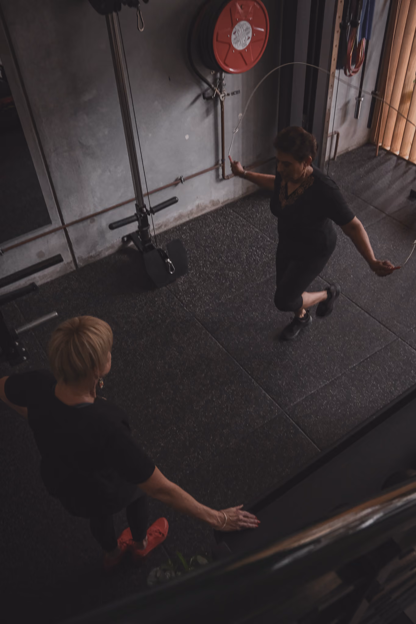 An elderly woman jumping rope in a gym while a trainer watches her performance.