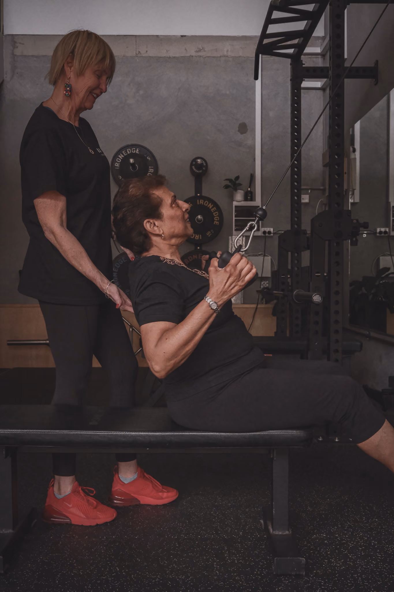 Older woman exercises with lat pulldown machine while another woman, possibly trainer, watches attentively.