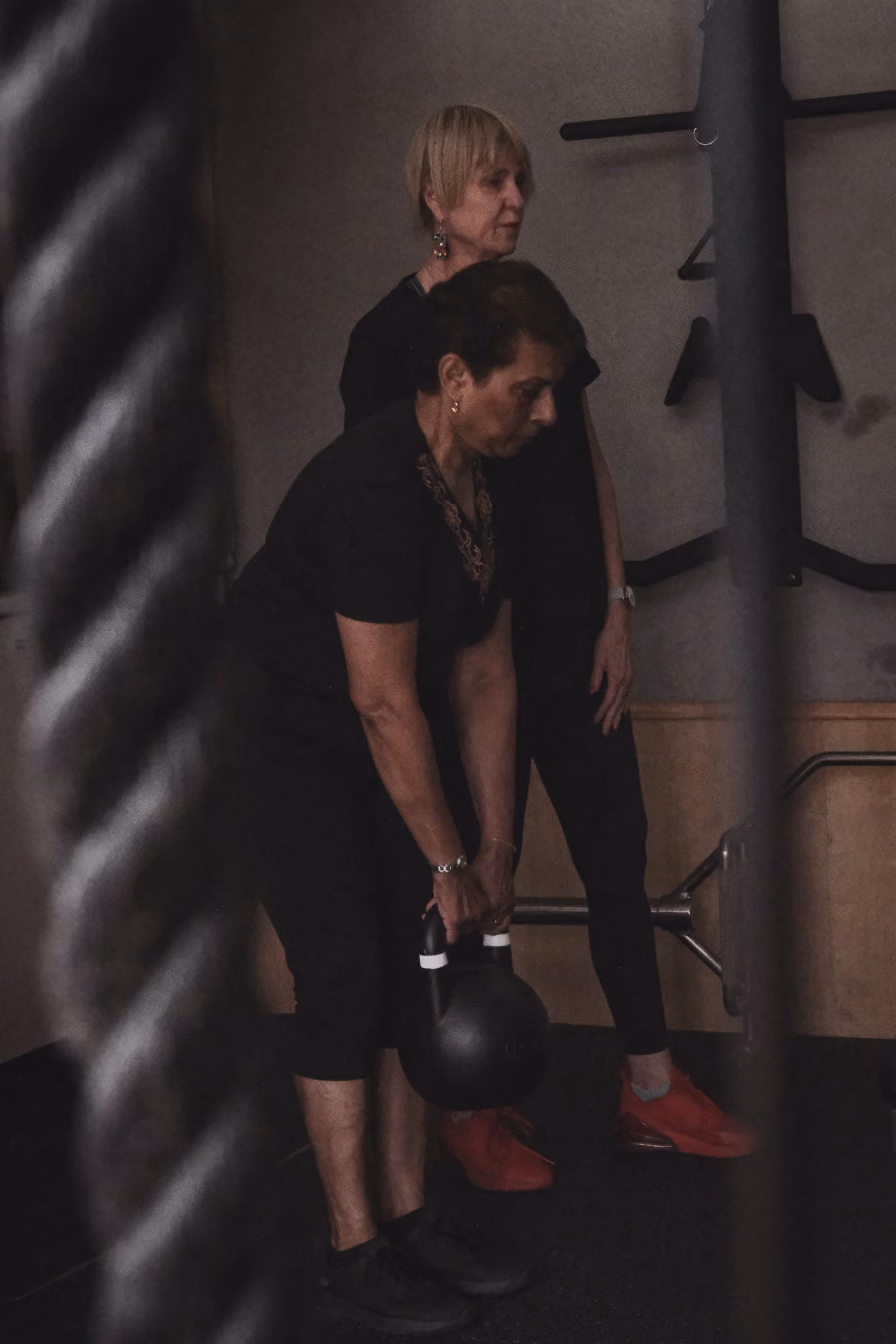 Two women in workout clothes in a gym, one lifting a kettlebell while the other watches.