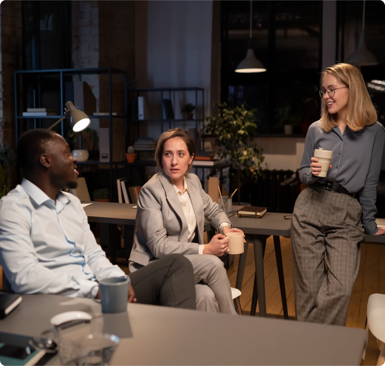 Three coworkers conversing in a modern office at night, each holding a coffee mug.