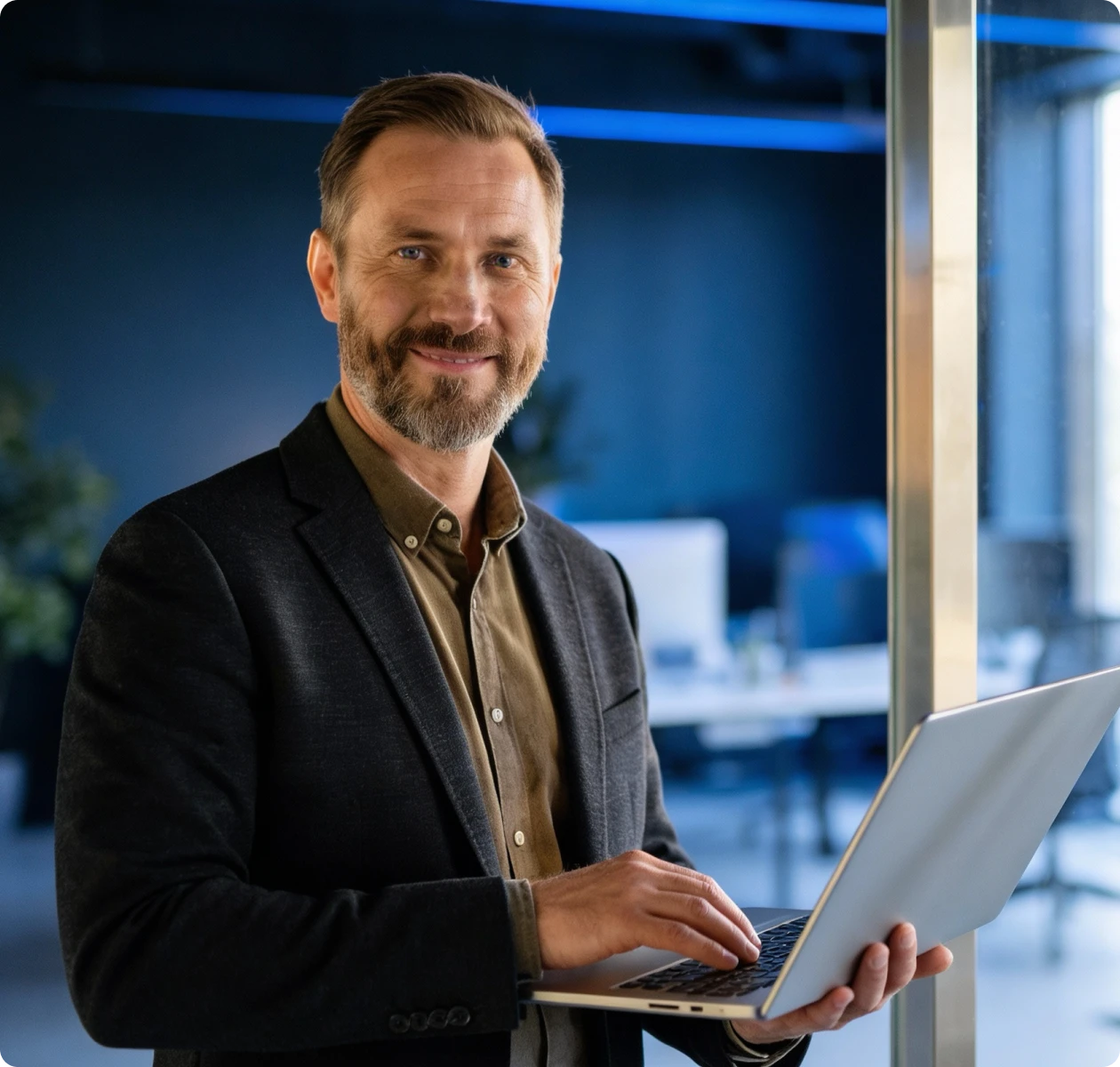 Smiling middle-aged man in a dark blazer holding and typing on a laptop in a modern office.