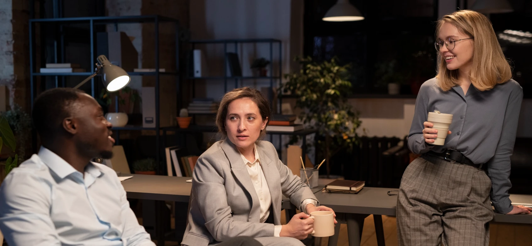 Three coworkers conversing in a modern office at night, each holding a coffee mug.