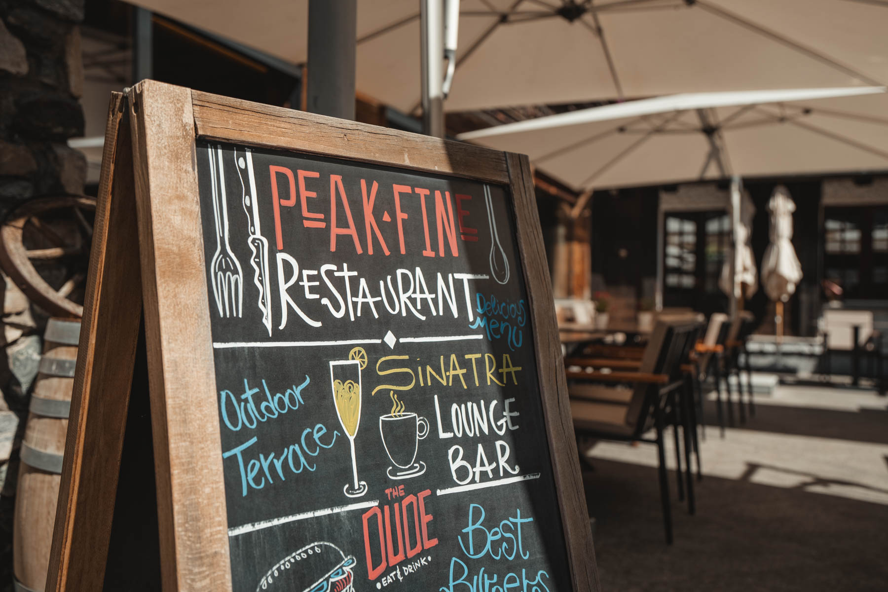 Ein hölzernes Kreidetafel-Schild im Aussenbereich des Alpen Resorts Zermatt präsentiert die gastronomischen Angebote: das Peak Fine Restaurant, die Sinatra Lounge Bar und «The Dude» für Burger, im Hintergrund ist die sonnige Aussenterrasse mit Sitzgelegenheiten und Sonnenschirmen zu sehen.