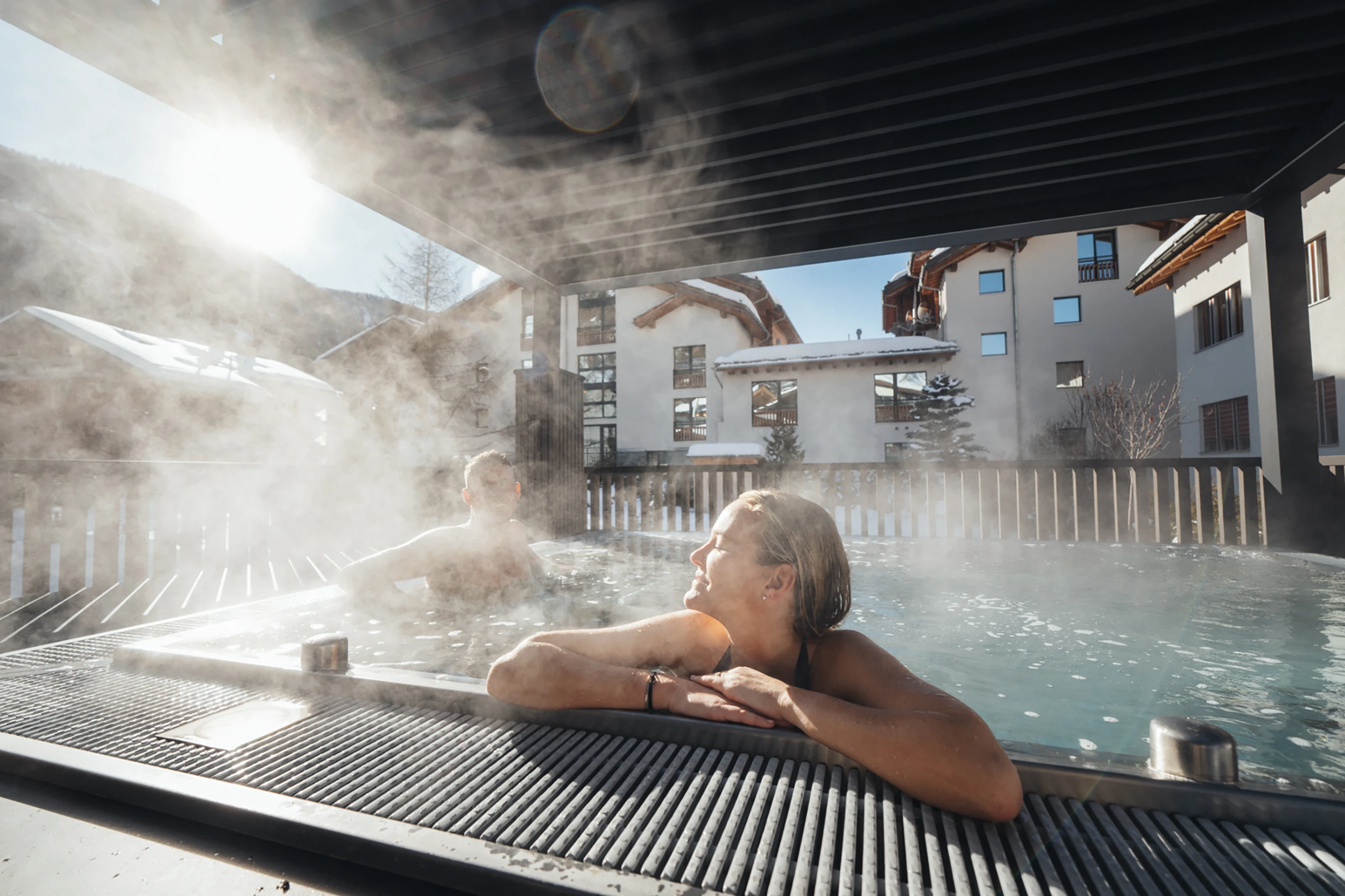 Ein Paar entspannt im dampfenden Aussen-Whirlpool des Alpen Resorts Zermatt bei strahlendem Sonnenschein, mit Blick auf die verschneiten Hotelgebäude und die umliegende Walliser Bergwelt.