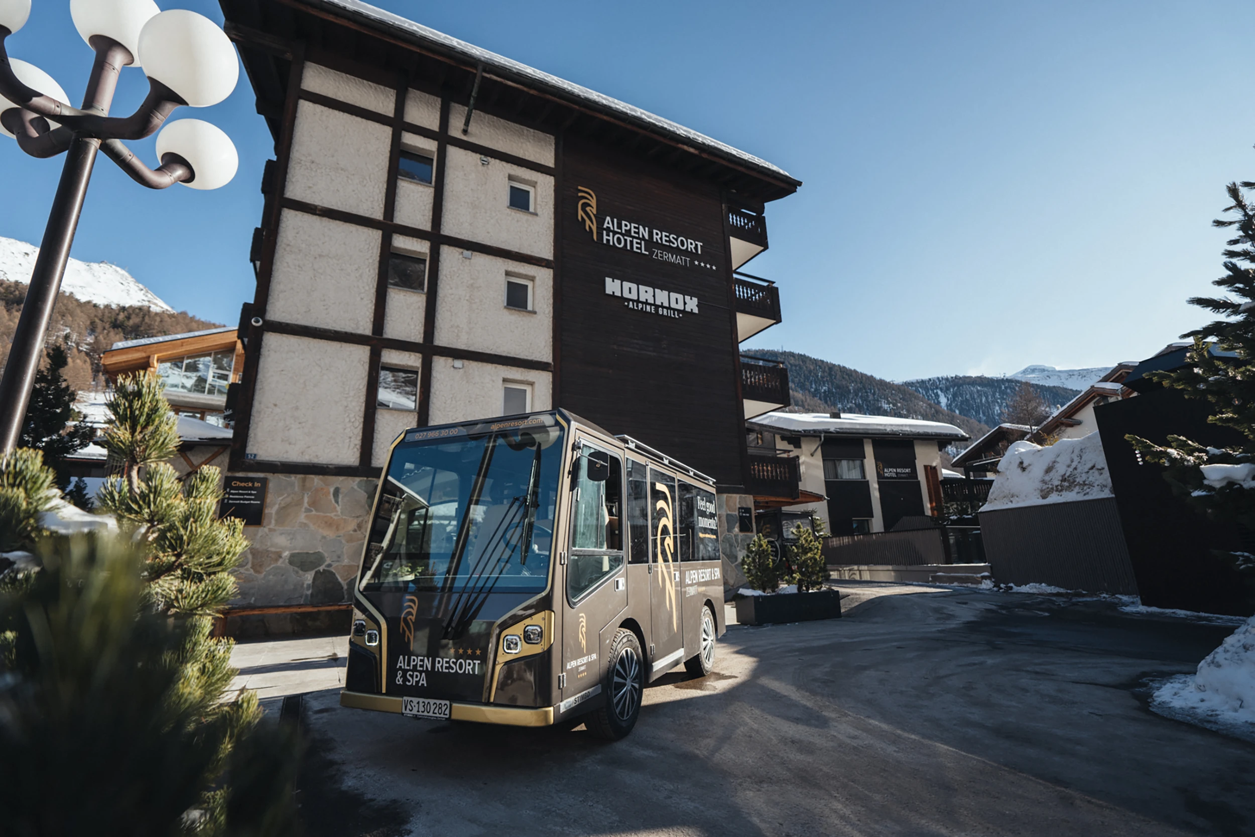 Das Elektro-Shuttle des Alpen Resorts Zermatt steht bereit vor der Hotelfassade mit den Logos des Hotels und des Hornox Alpine Grills, um Gäste im autofreien Zermatt komfortabel zu transportieren.