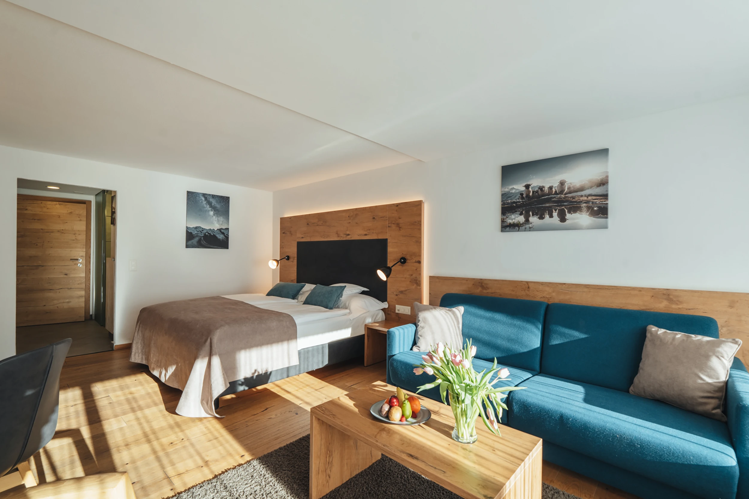 Ein modern eingerichtetes Hotelzimmer im Alpen Resort Zermatt mit hellem Parkettboden, einem Doppelbett mit Holz-Kopfteil, einem blauen Sofa und einem Couchtisch mit frischen Blumen und Früchten.