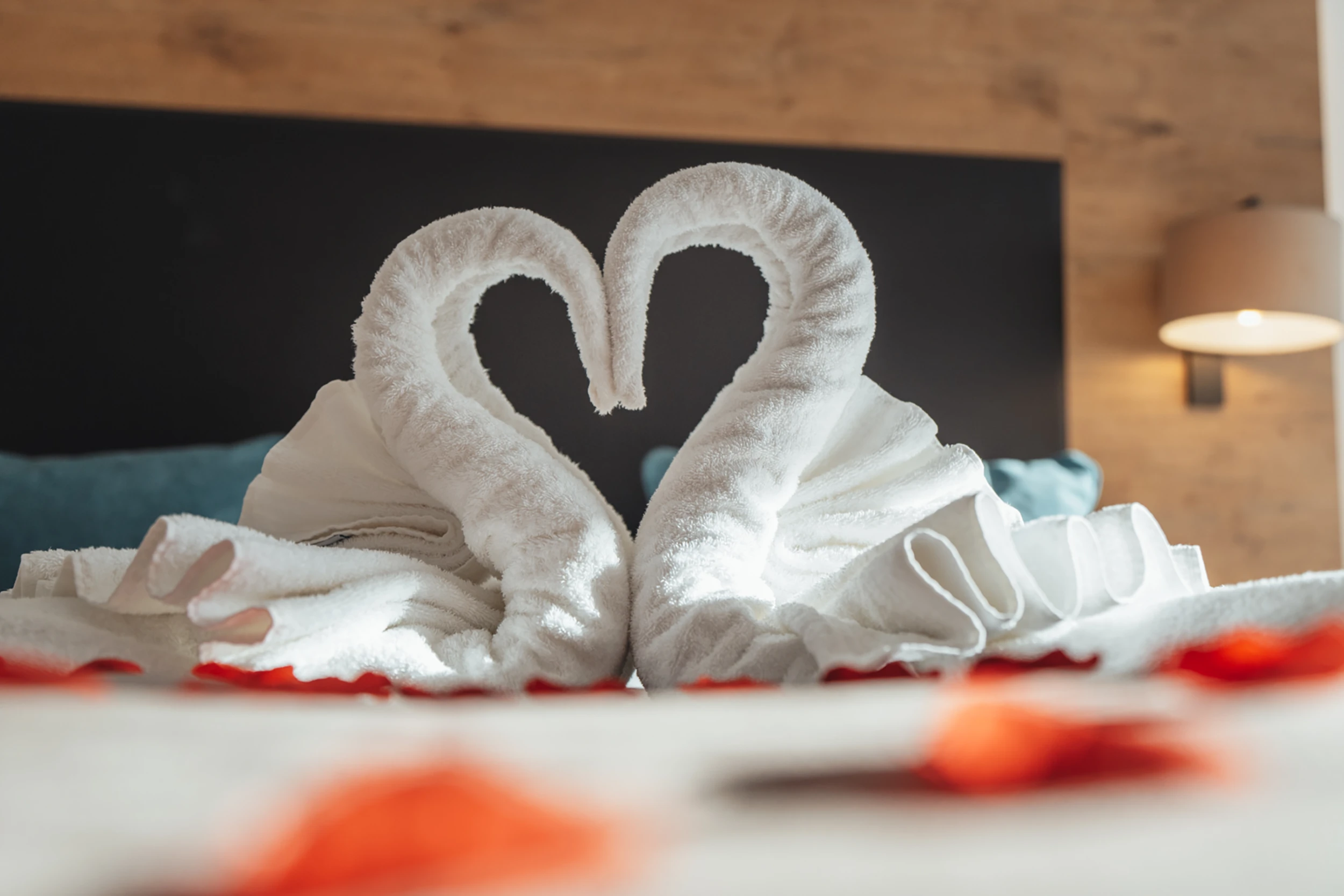 Zwei kunstvoll zu Schwänen geformte weisse Handtücher bilden ein Herz auf einem Hotelbett im Alpen Resort Zermatt, dekoriert mit roten Rosenblüten für einen romantischen Empfang.