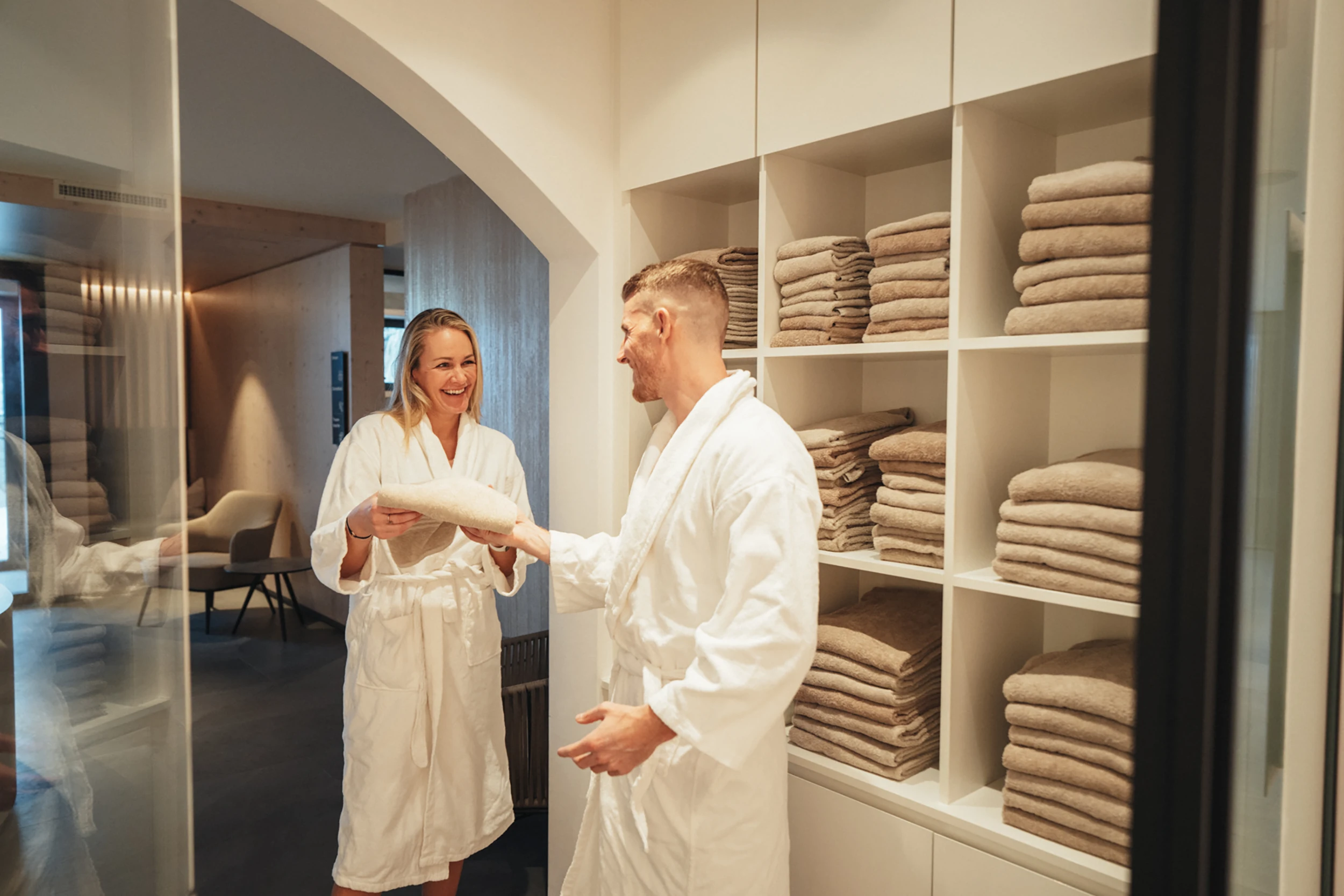 Ein lächelndes Paar in weissen Bademänteln bereitet sich im Wellnessbereich des Alpen Resorts Zermatt auf einen entspannten Tag vor, während sie sich bei einem Regal mit frisch zusammengelegten Handtüchern unterhalten.