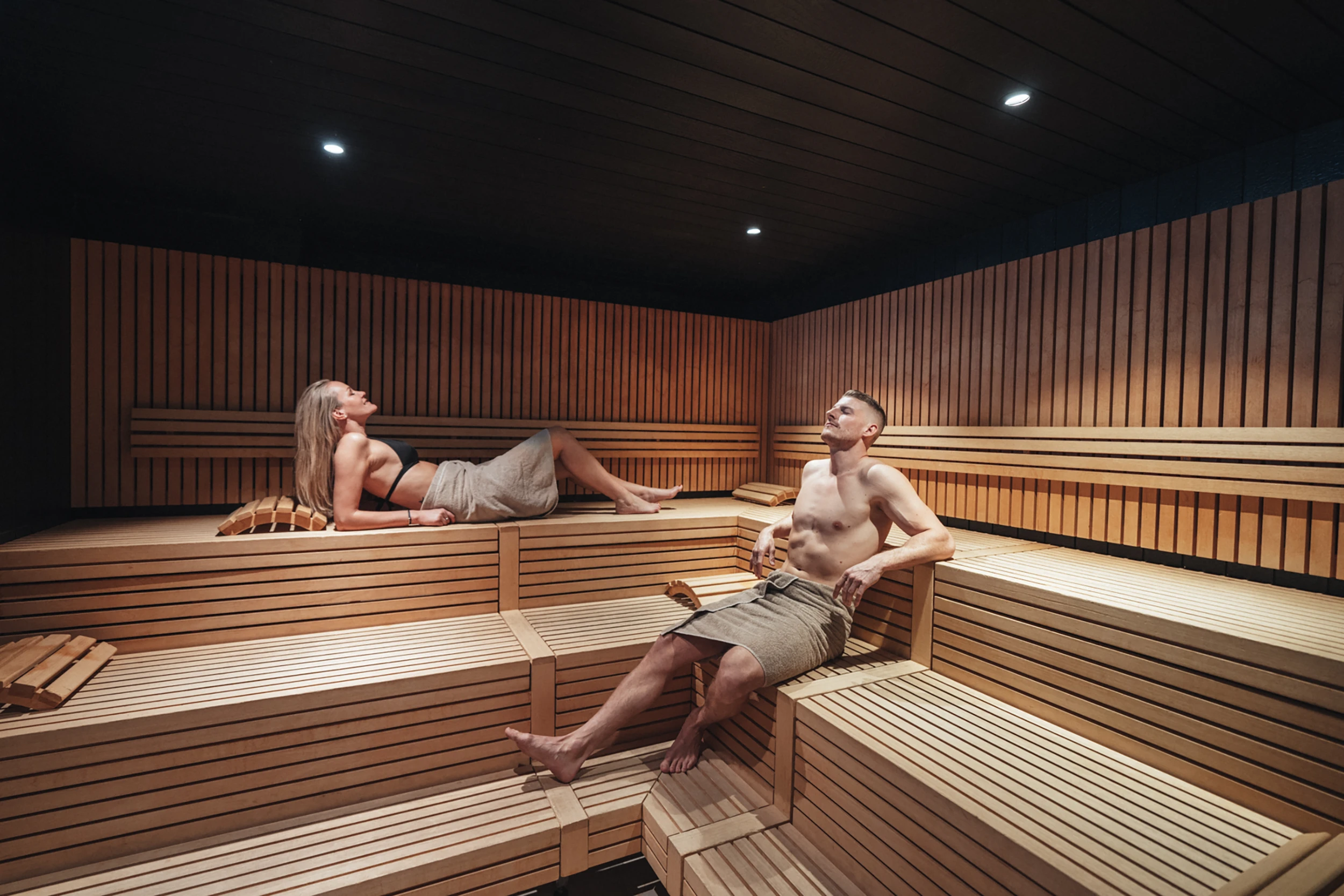 Ein Paar entspannt in der grosszügigen, modern gestalteten Holzsauna des Alpen Resorts Zermatt und geniesst die wohltuende Wärme im Wellnessbereich.
