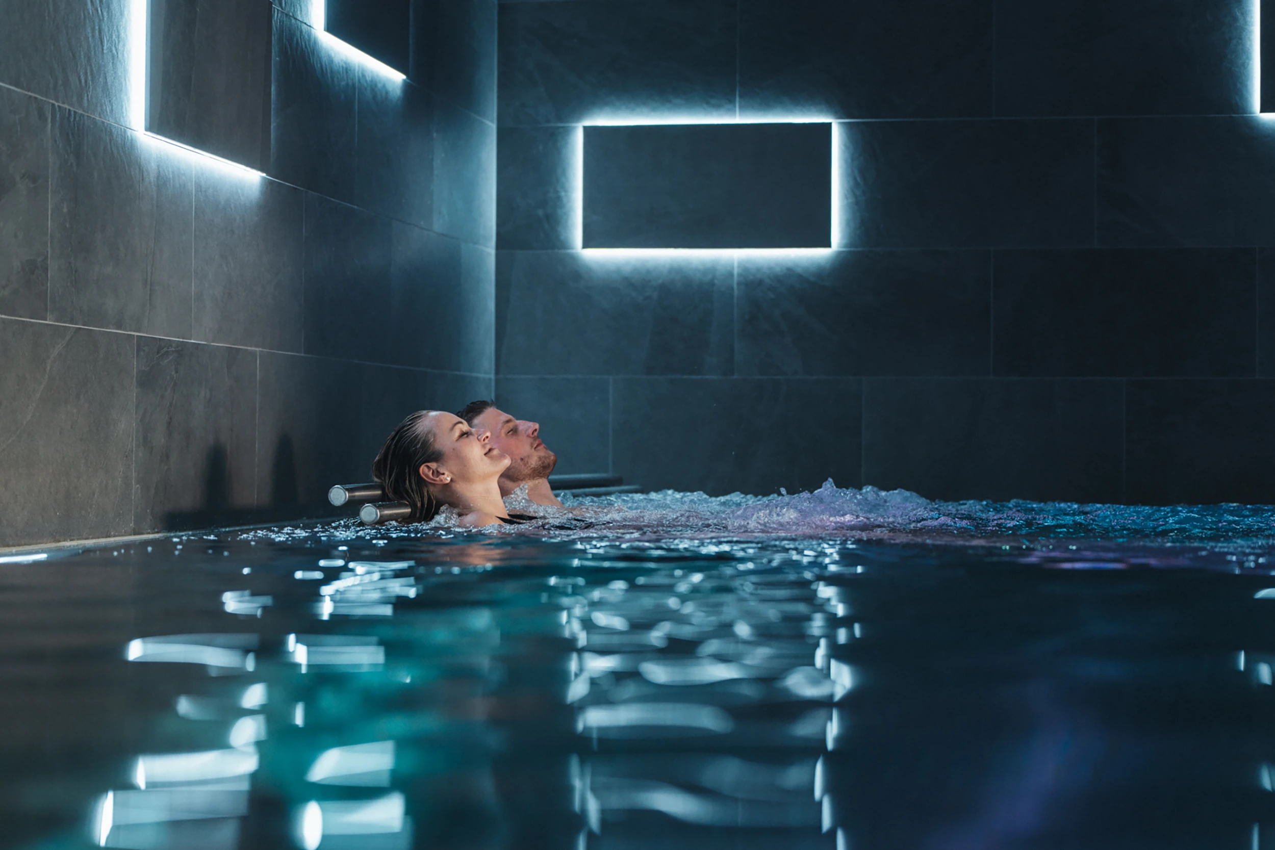 Ein Paar entspannt in den Sprudelliegen des stilvoll beleuchteten Indoor-Pools im Alpen Resort Zermatt, umgeben von dunklen Steinwänden mit modernen, quadratischen Lichtelementen.