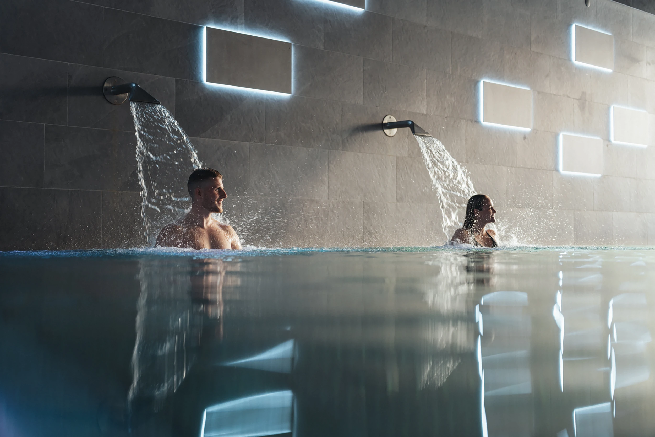 Ein Paar geniesst die Nackenduschen im modernen Indoor-Pool des Alpen Resorts Zermatt, während das Wasser vor einer stilvoll beleuchteten Steinwand in das Becken sprudelt.