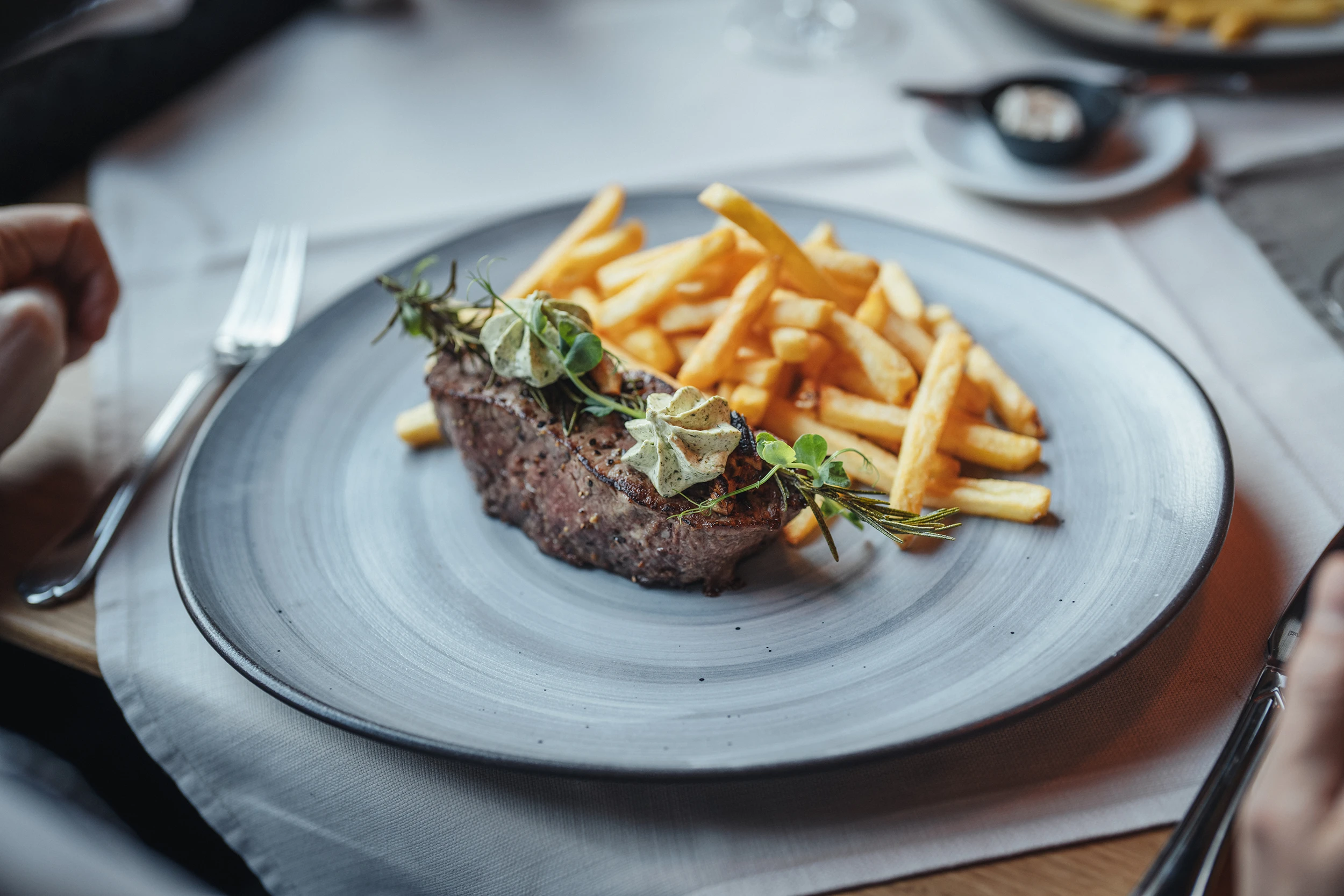 Ein perfekt gebratenes Steak, garniert mit Kräuterbutter und frischem Rosmarin, serviert mit knusprigen Pommes Frites auf einem modernen grauen Teller im Restaurant PeakFine.