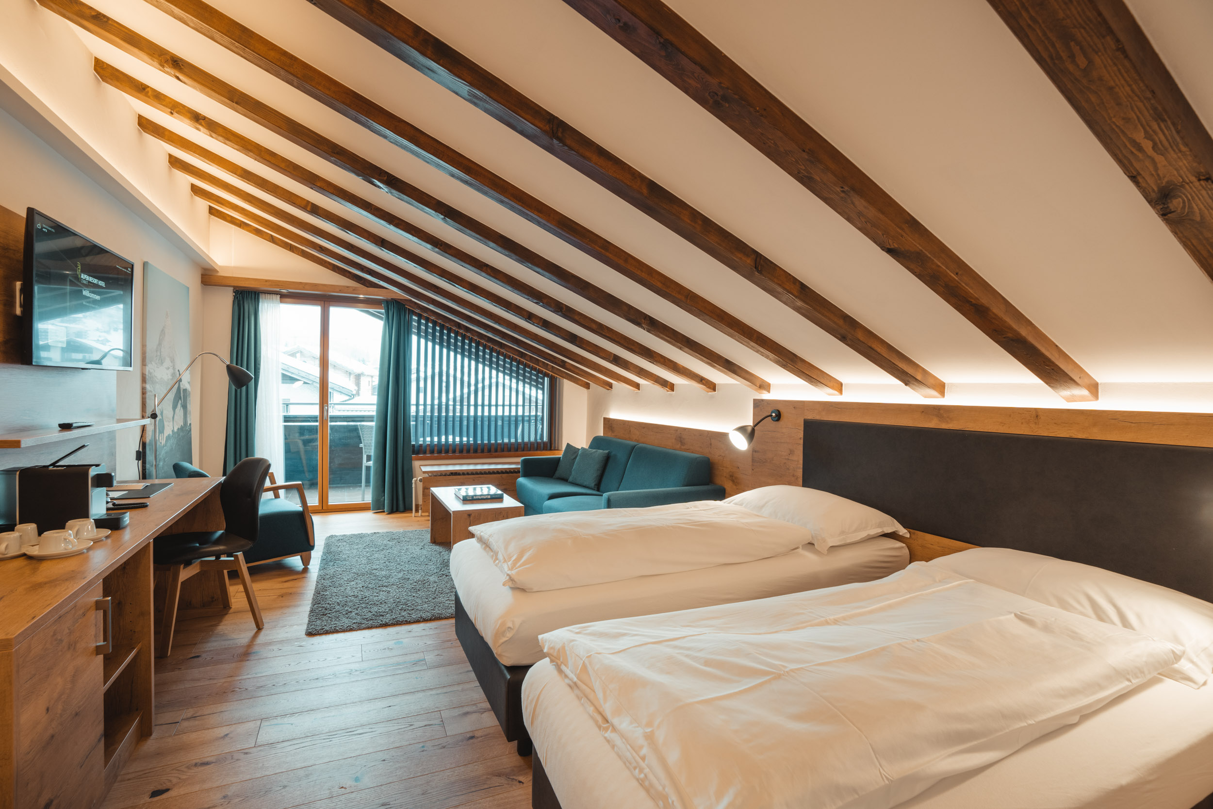 Ein gemütliches Hotelzimmer im Alpen Resort Zermatt mit markanten Sichtbalken an der Dachschräge, zwei Einzelbetten, einer Sitzecke mit blauem Sofa und einem Schreibtisch aus hellem Holz.