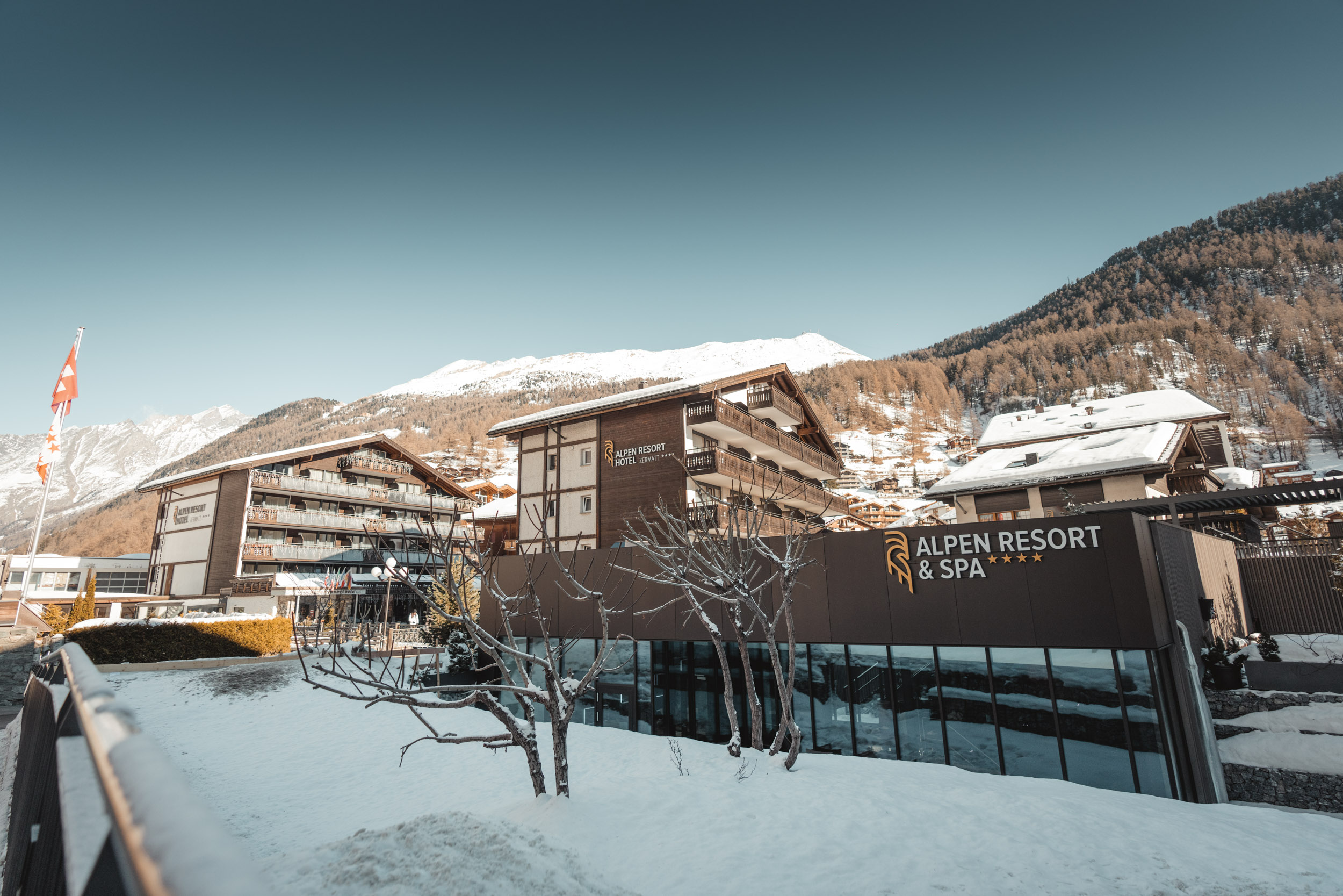 Die Aussenansicht des Alpen Resorts & Spa in Zermatt im Winter, mit den charakteristischen Hotelgebäuden aus Holz und Stein vor einer verschneiten Bergkulisse und blauem Himmel.