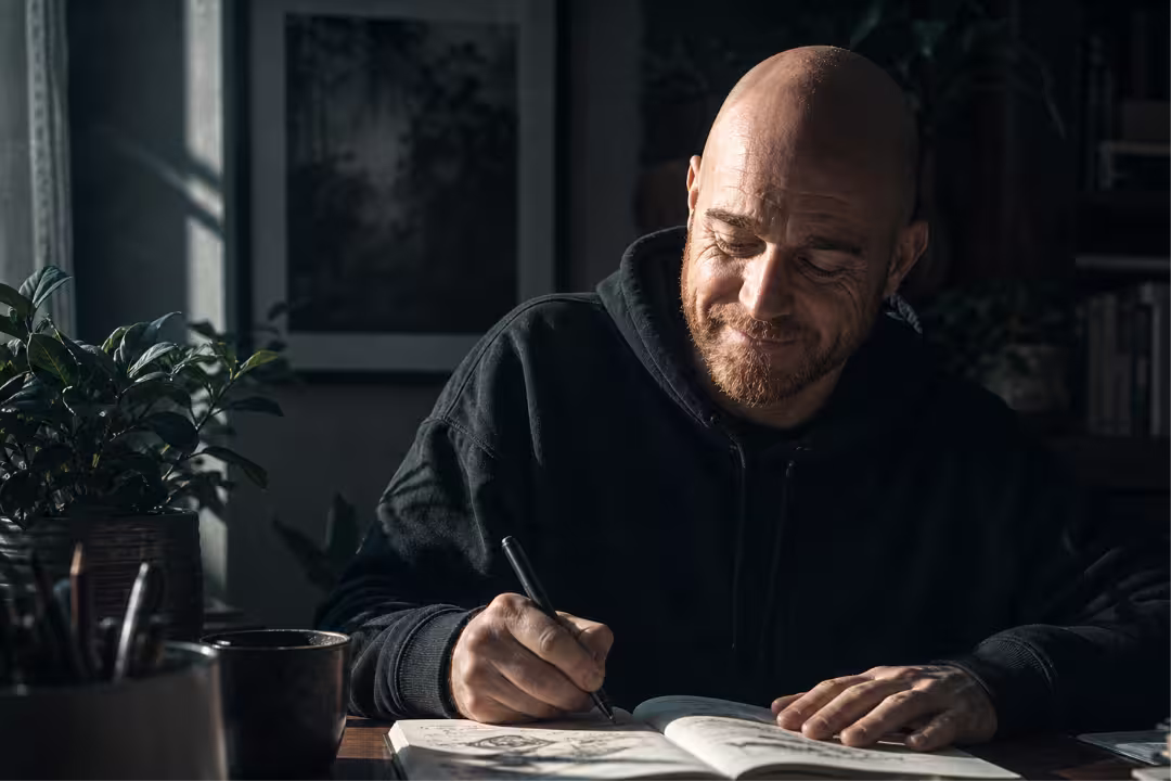 Smiling designer sketching ideas in a notebook at a sunlit wooden desk.
