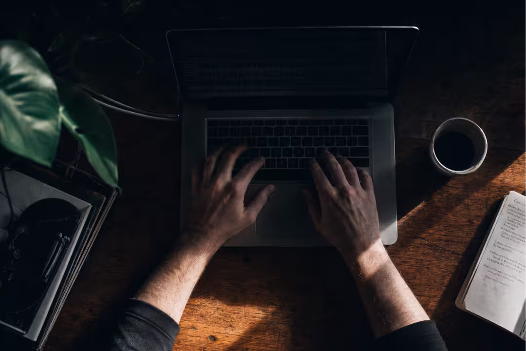 Top-down view of hands typing on a laptop at a rustic wooden desk with coffee and notes.