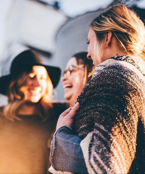 Three women outdoors smiling and laughing together warmly in the sunlight.