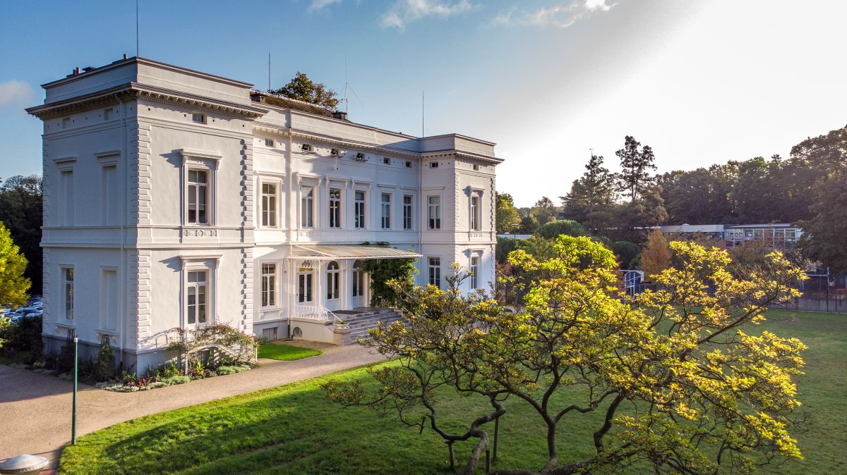A large white classical building surrounded by green lawn and trees under a partly cloudy sky.