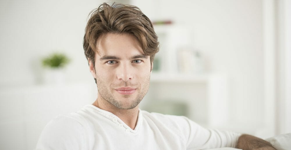 Young man with brown hair and stubble wearing a white shirt, sitting on a couch with a blurred background of a plant and bookshelf.